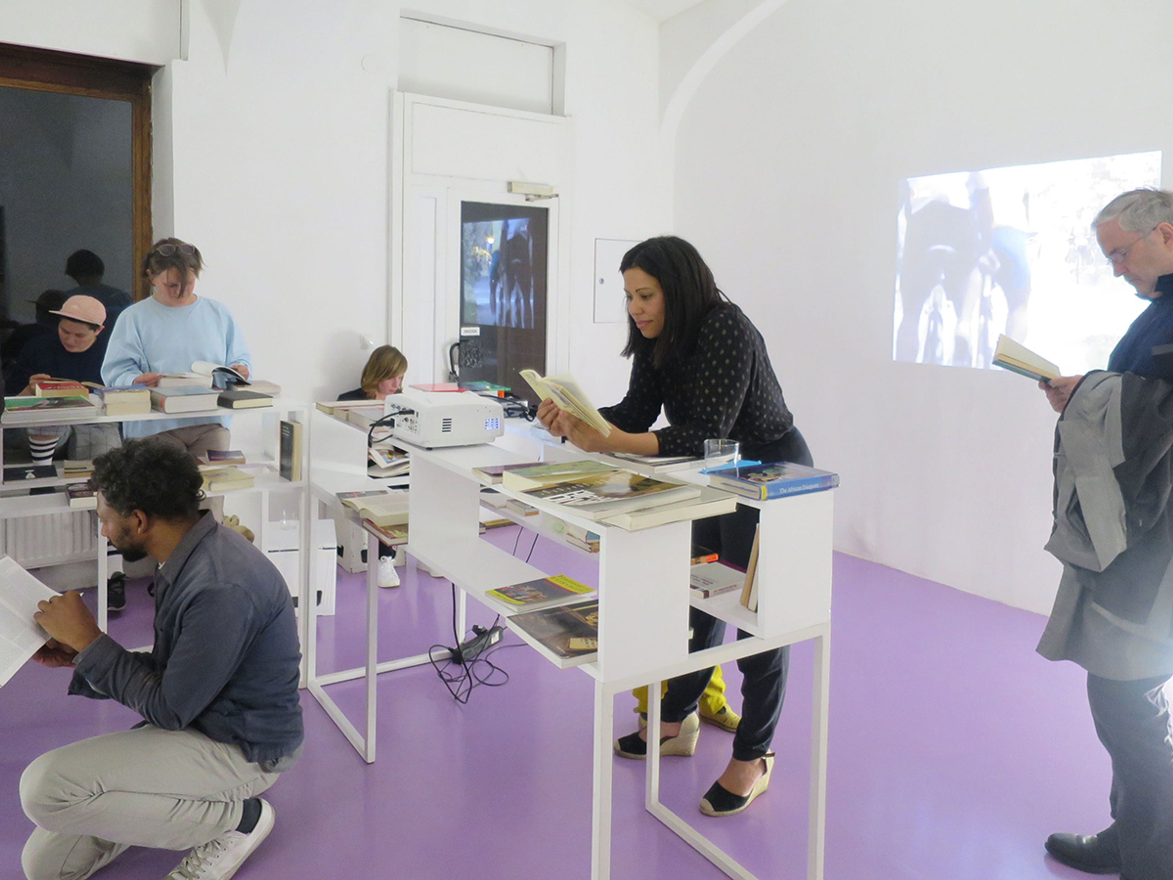 People reading books around white display tables in a modern room with a lavender floor and a projected image on the wall.