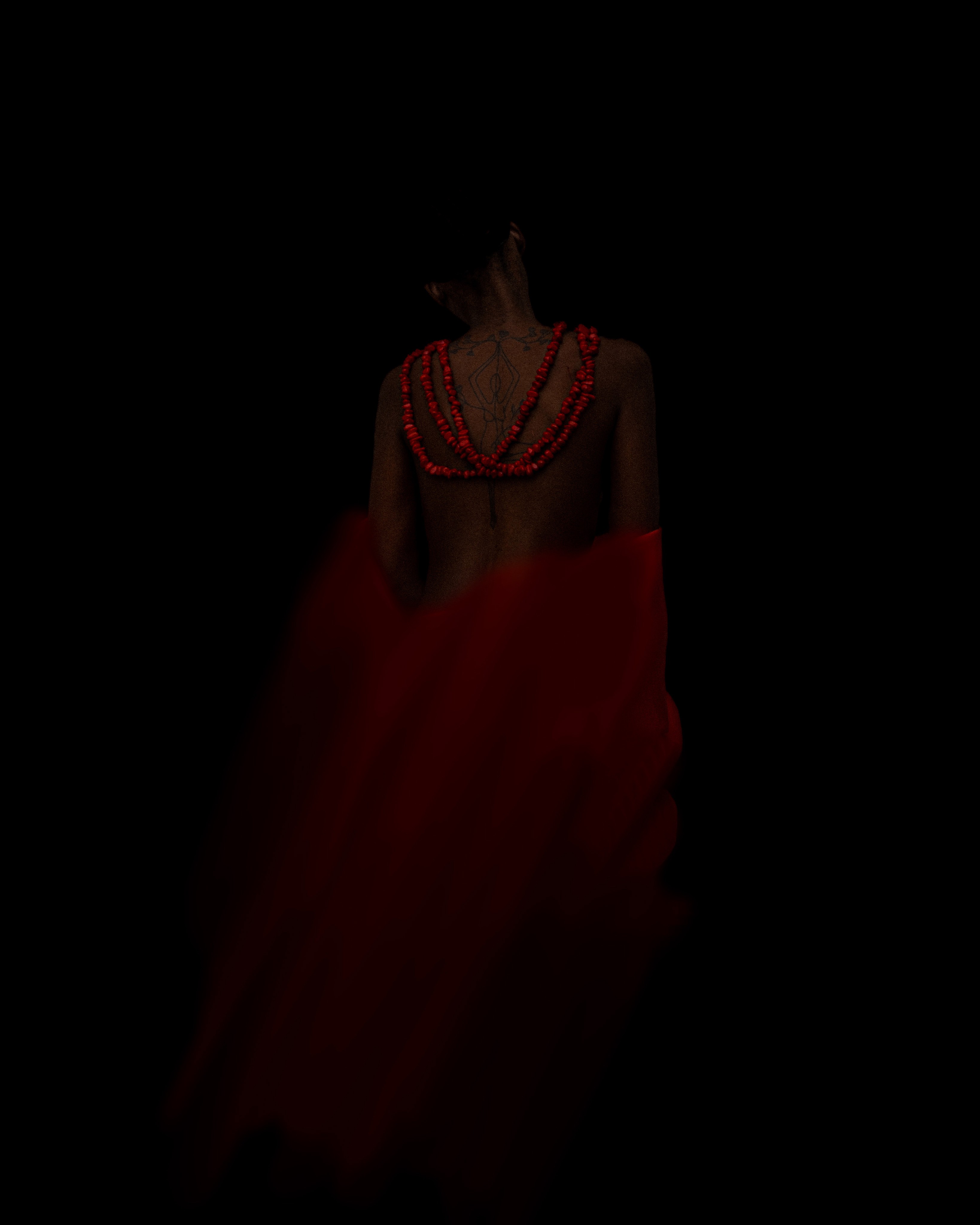 A person's back adorned with red beads and flowing red fabric, set against a dark background.