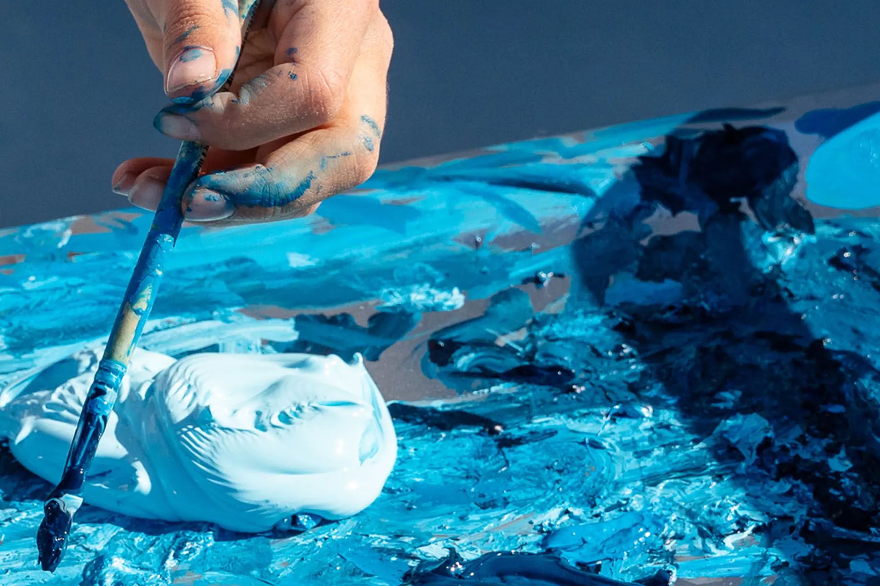 A hand with blue paint holds a brush, mixing various shades of blue paint on a palette.
