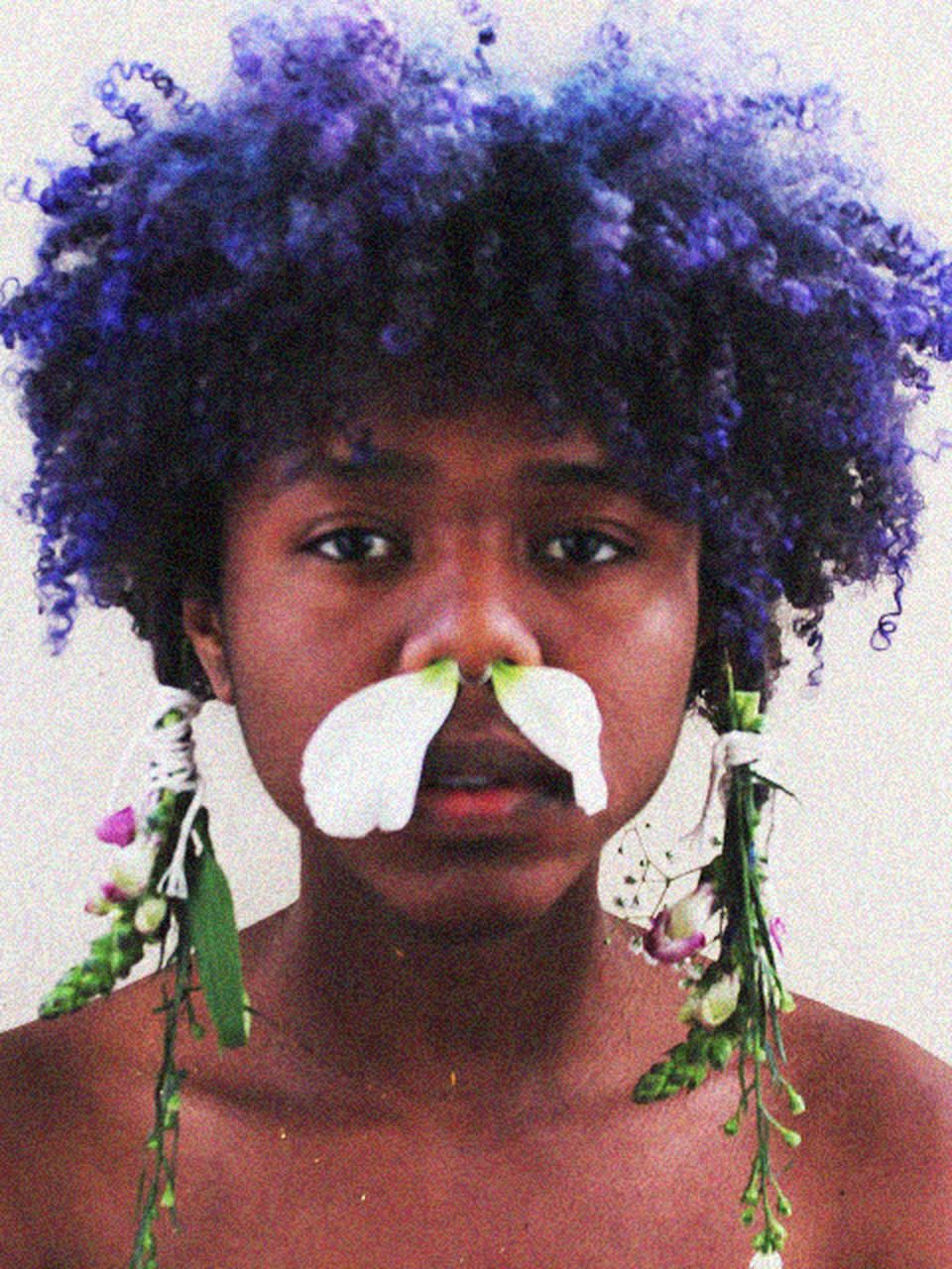 Portrait of a person with purple curly hair, a white petal mustache, and flowers draped from their hair.
