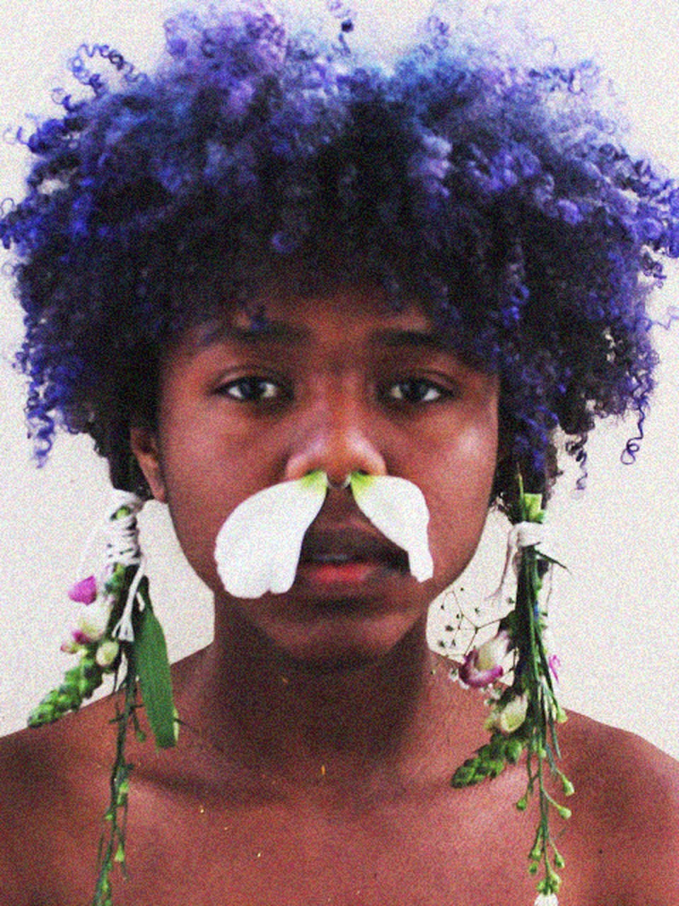 Portrait of a person with purple curly hair, a white petal mustache, and flowers draped from their hair.
