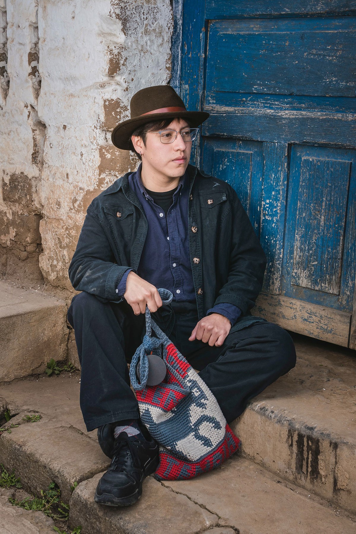 A young man with glasses and a hat sits on stone steps, holding a colorful woven bag, next to a weathered wall and a blue door.