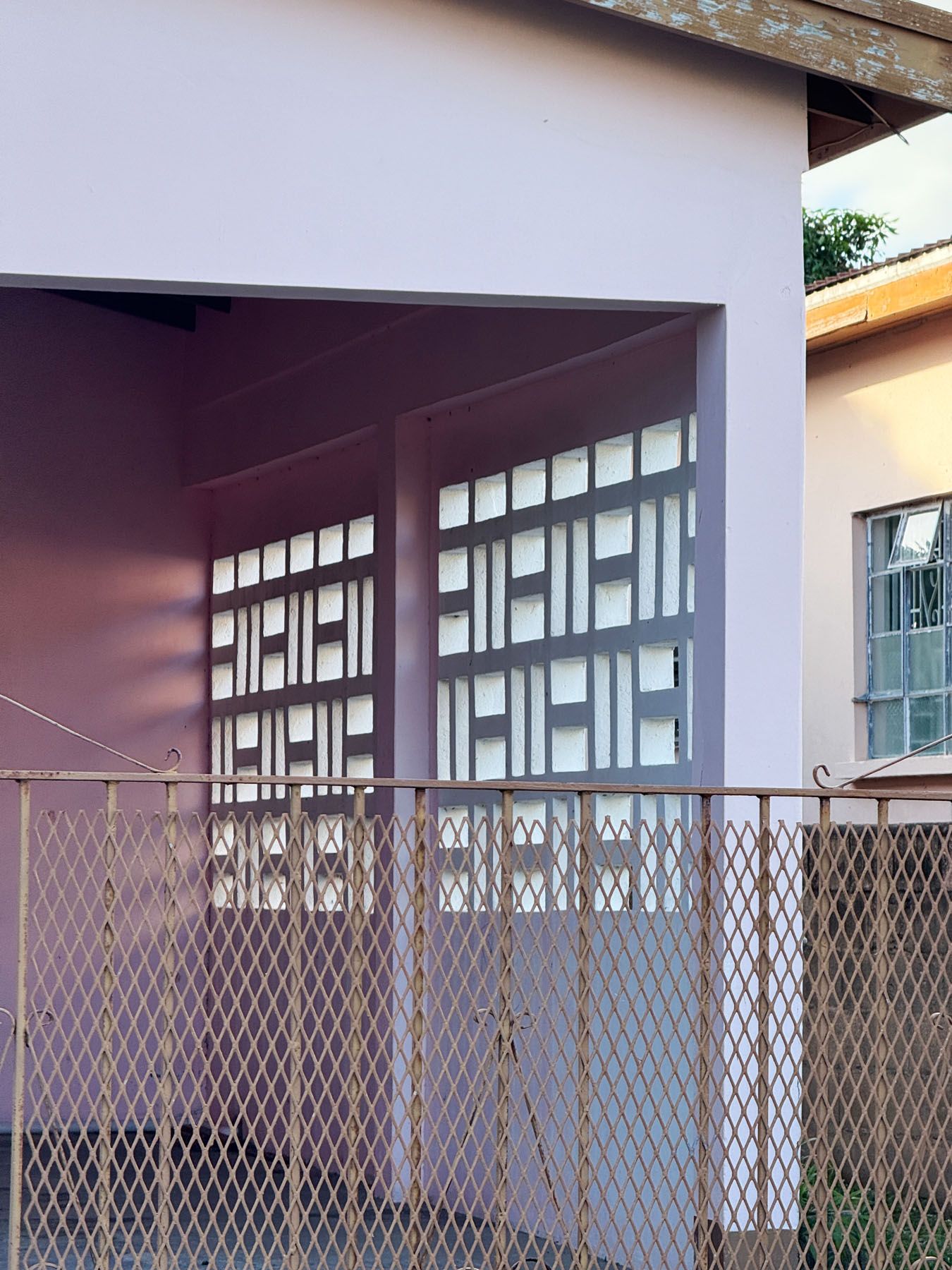A light purple building with a recessed porch featuring decorative white screens, behind a rusty metal mesh fence.