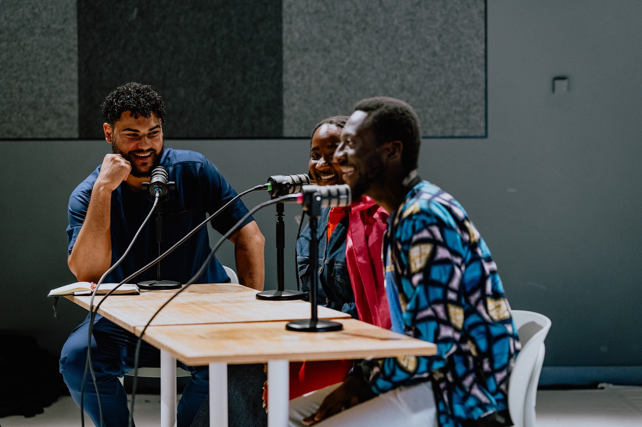 Three smiling people sit at a table with microphones, recording a podcast.