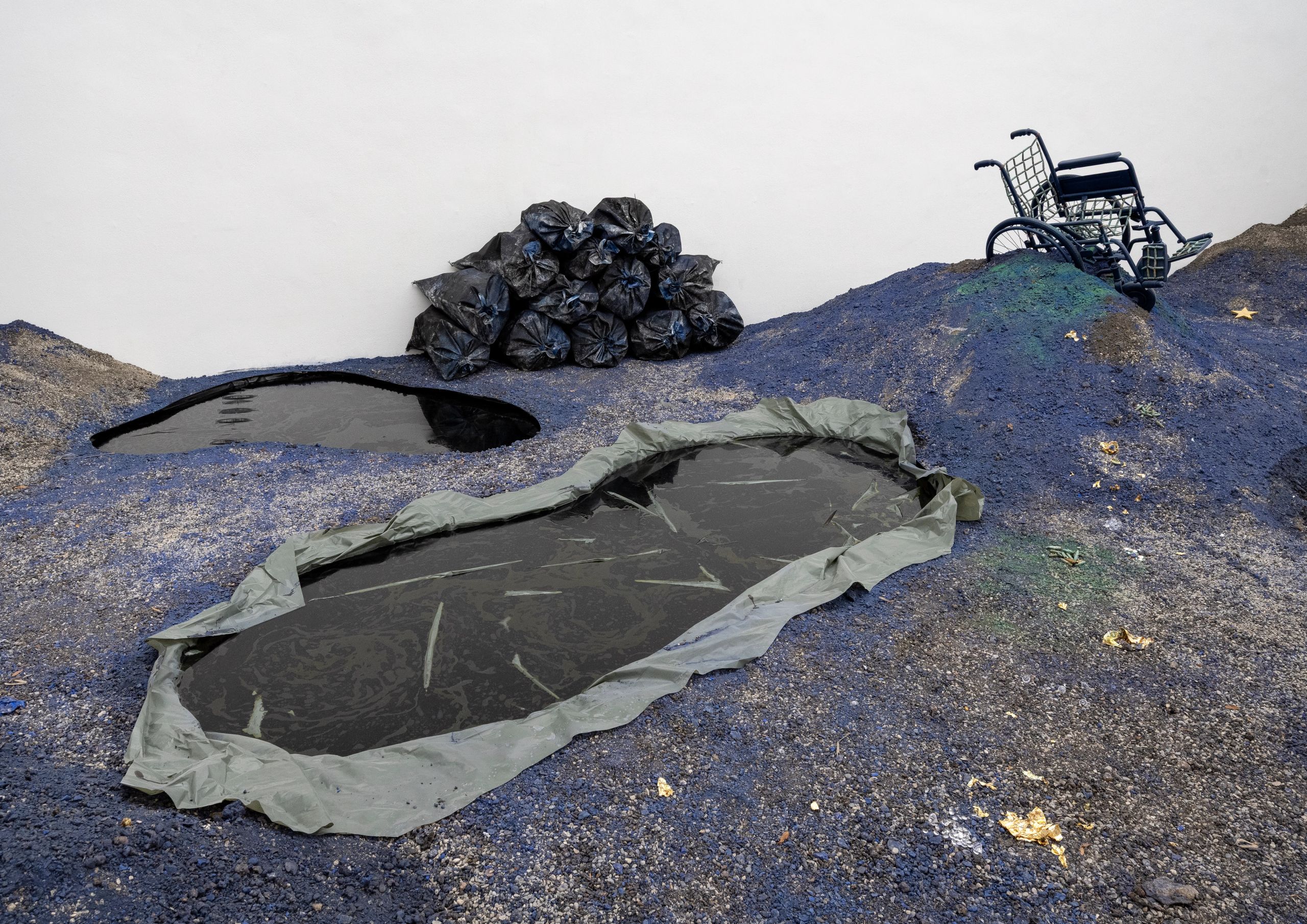 An art installation featuring blue-grey textured ground with two dark liquid pools, a pile of trash bags, and an overturned black wheelchair.