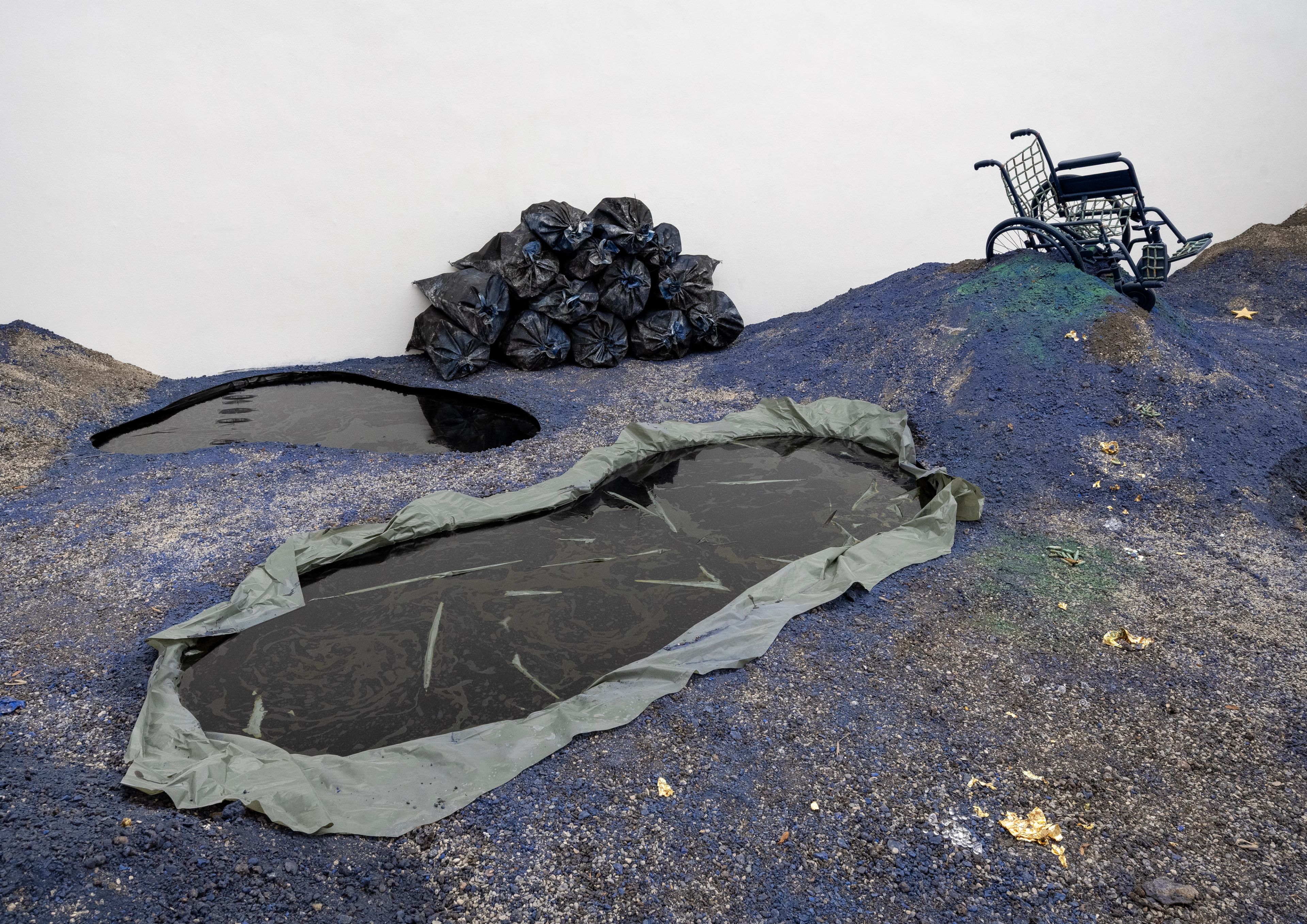 An art installation featuring blue-grey textured ground with two dark liquid pools, a pile of trash bags, and an overturned black wheelchair.