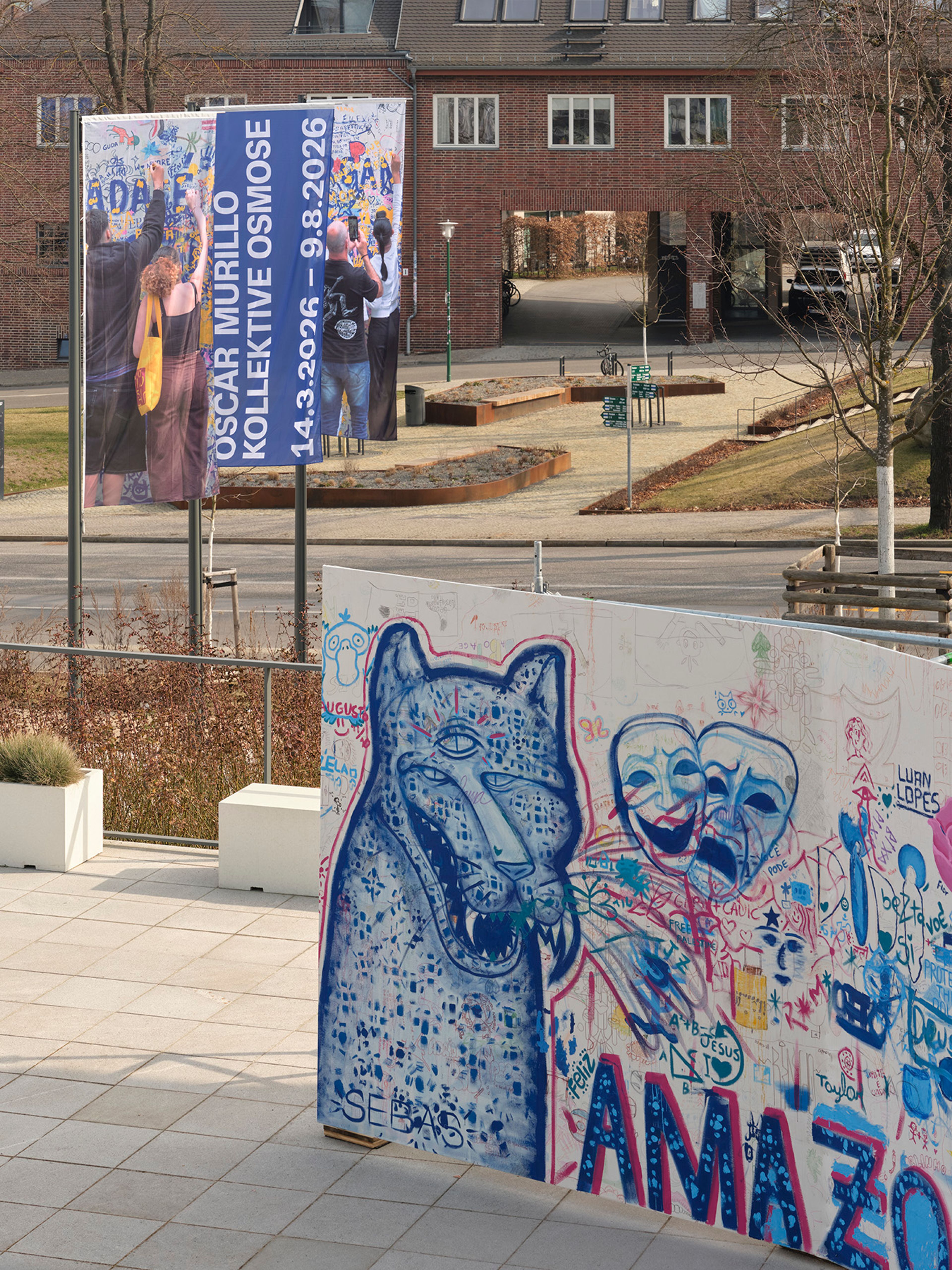 A large exhibition banner stands next to a graffiti-covered wall featuring a blue wolf and two theatrical masks, with a brick building in the background.