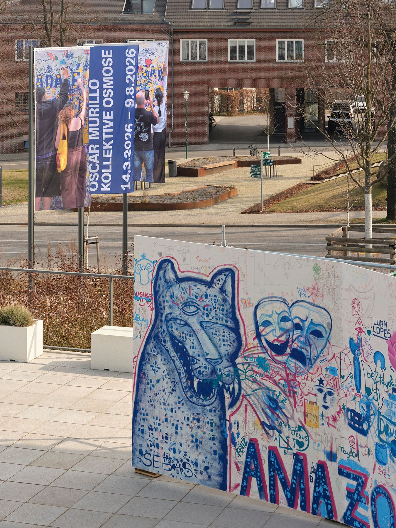 A large exhibition banner stands next to a graffiti-covered wall featuring a blue wolf and two theatrical masks, with a brick building in the background.