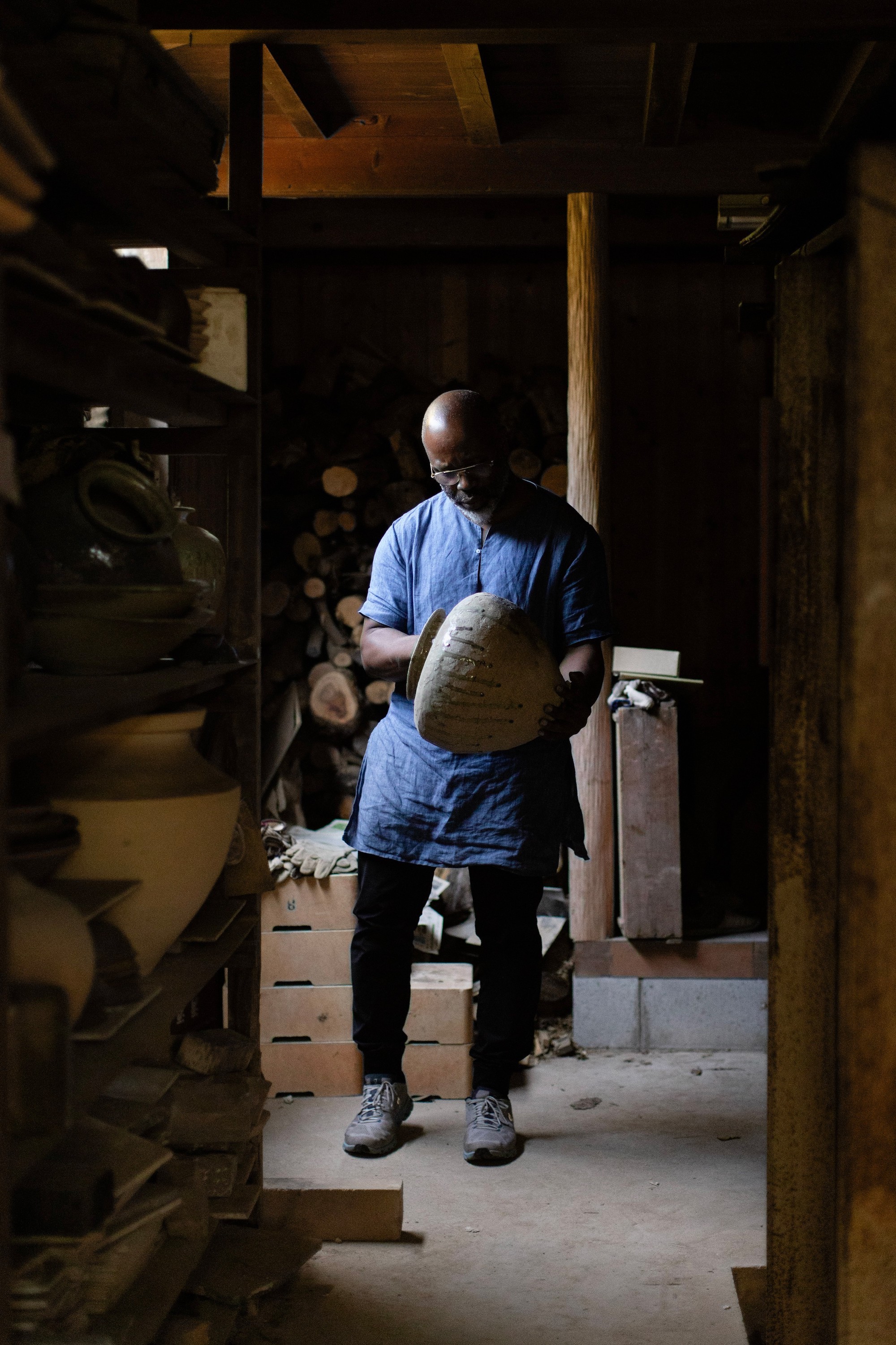 A man in a blue tunic holds a light-colored ceramic vessel in a dimly lit, rustic workshop with pottery on shelves and firewood in the background.