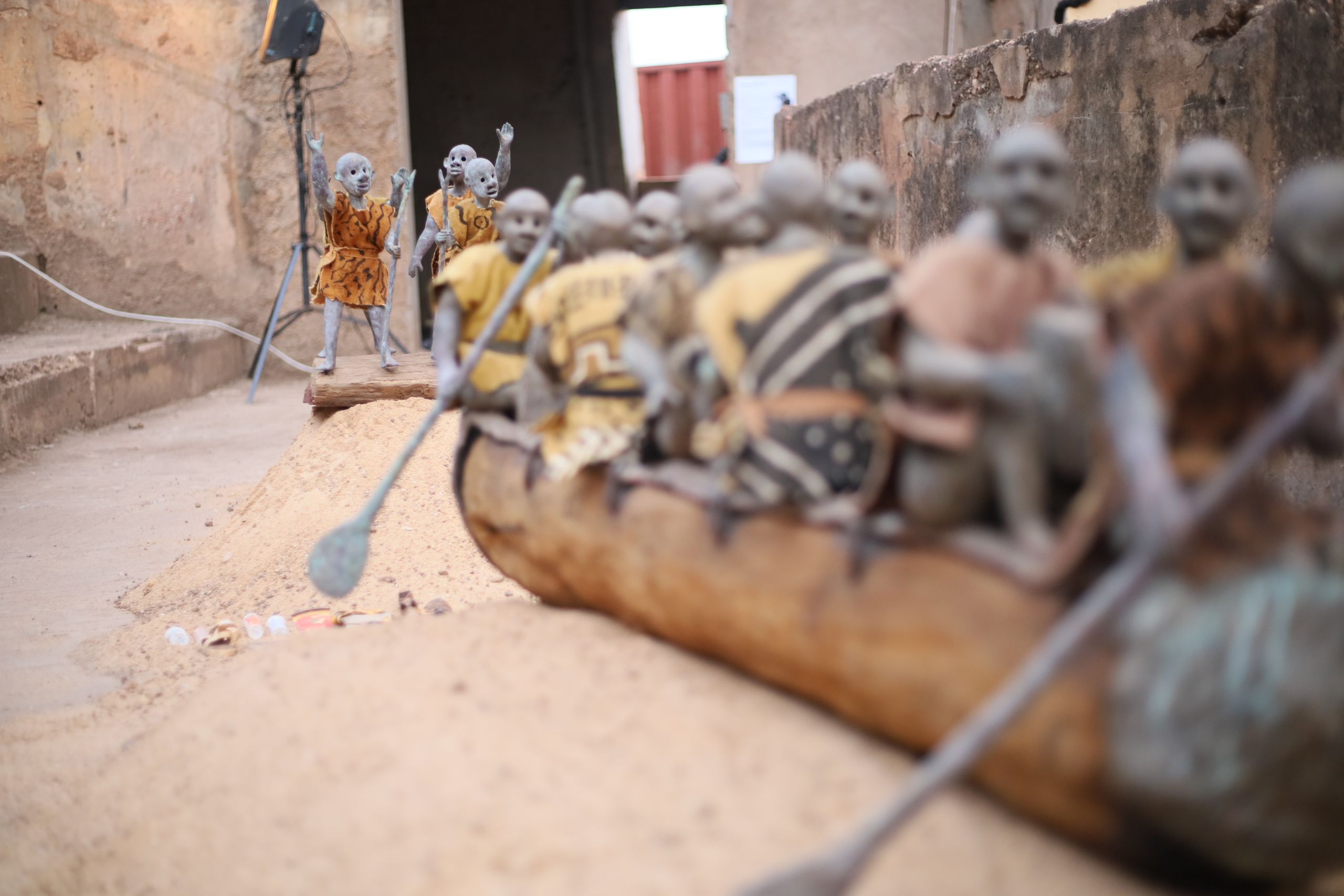 A diorama depicts traditional figures in a wooden canoe on a sandy bank, with more figures standing behind them; a studio light is visible in the background.
