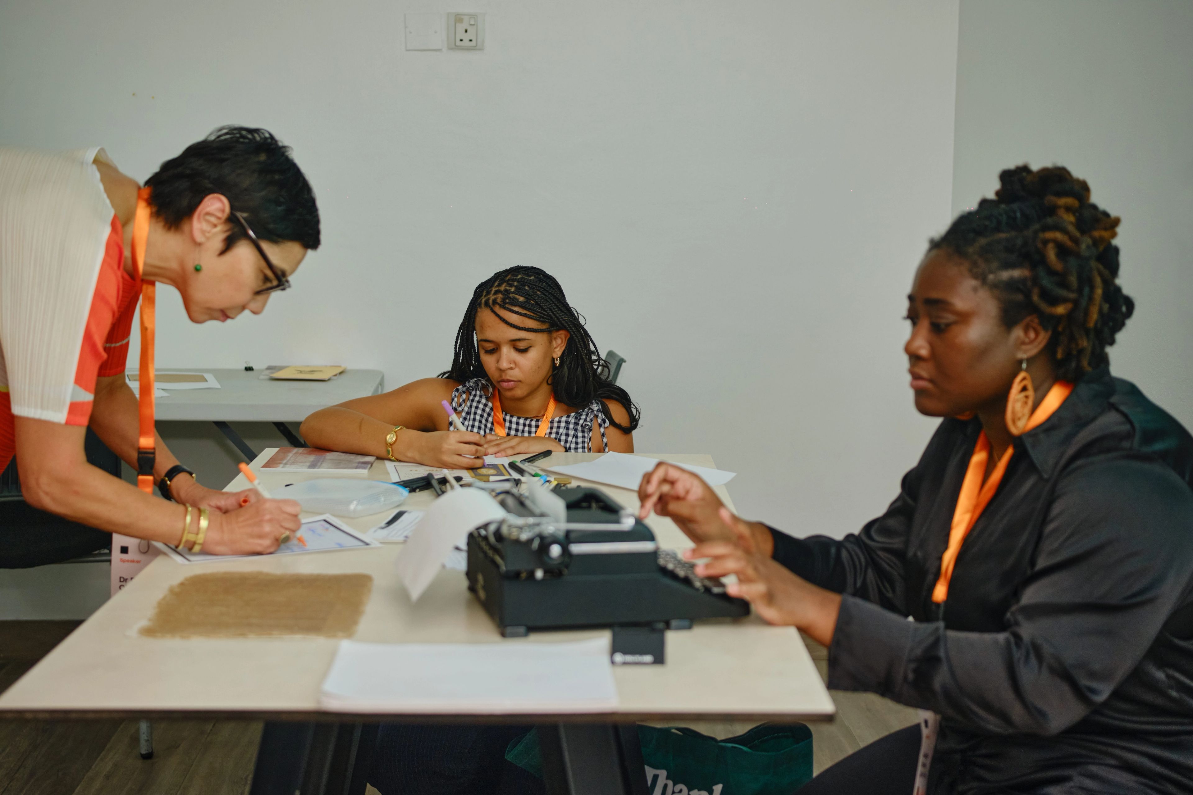 Three women at a table, one writing with a pen, another drawing with a marker, and a third typing on a vintage typewriter.
