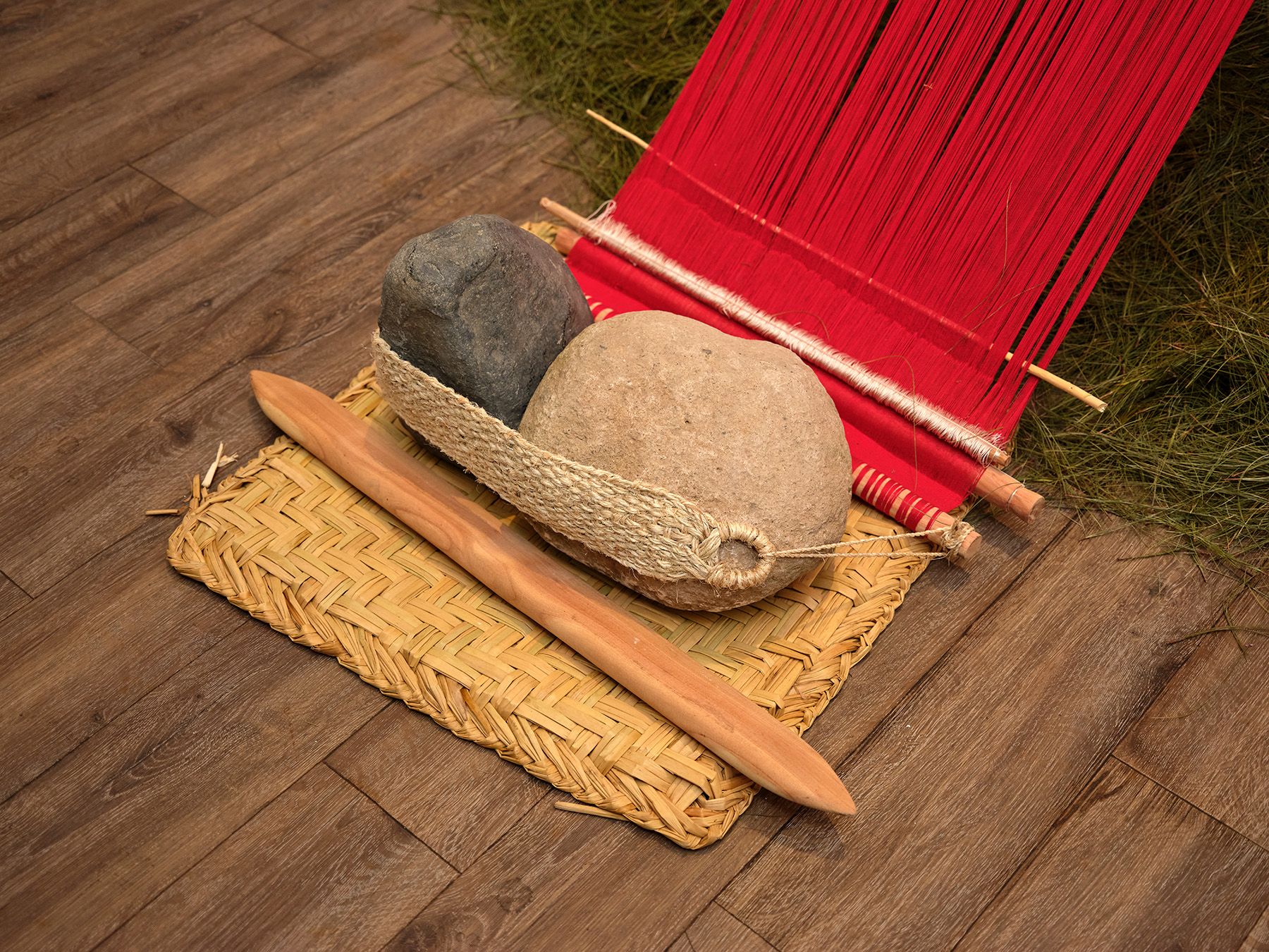 Loom with red threads, two stones in a woven basket, and a wooden tool on a woven mat.