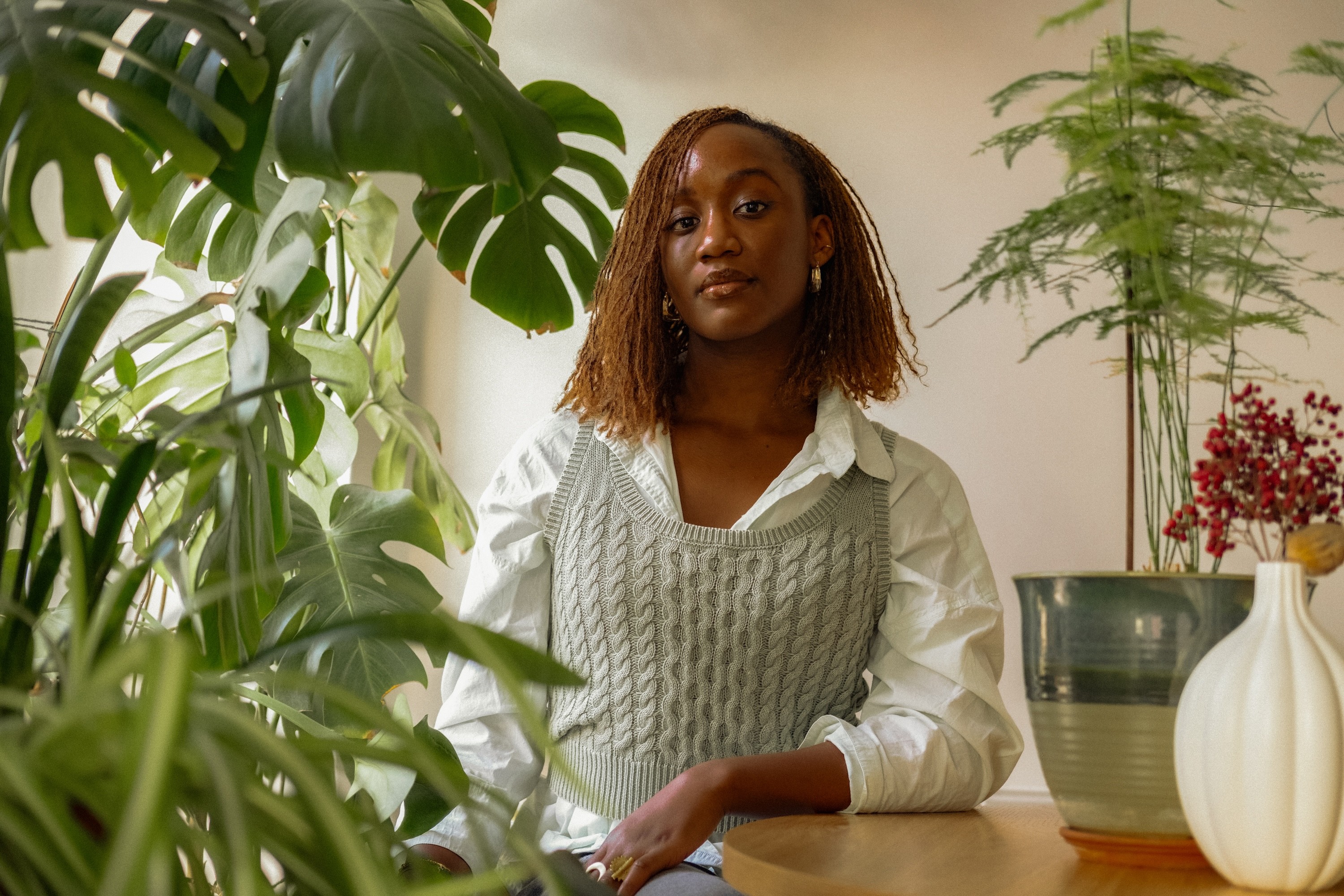 A young Black woman with brown dreadlocks sits at a table surrounded by plants, wearing a white shirt and gray sweater vest, looking at the camera.