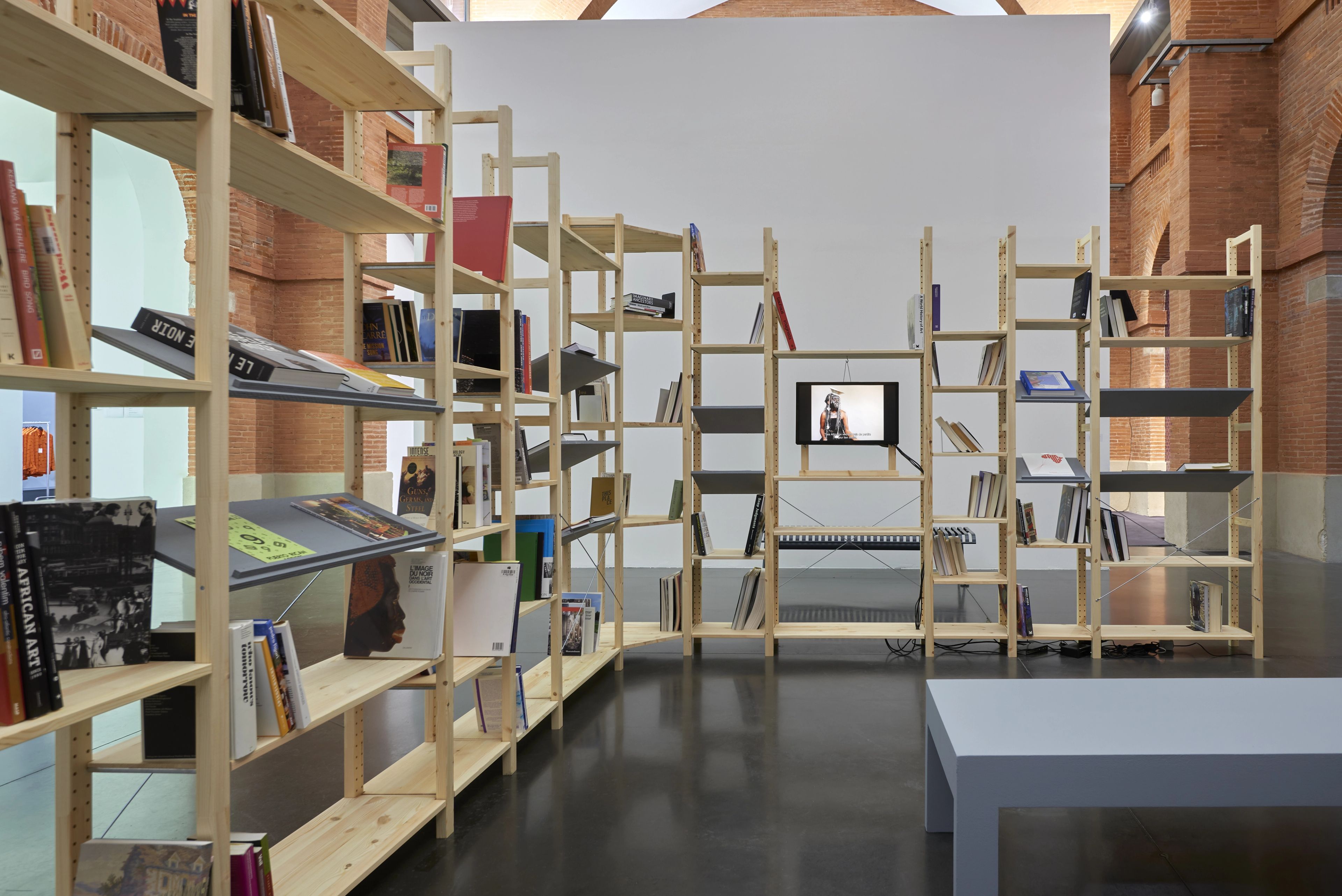 L-shaped wooden bookshelves displaying books and a video portrait, within an exhibition space with exposed brick.