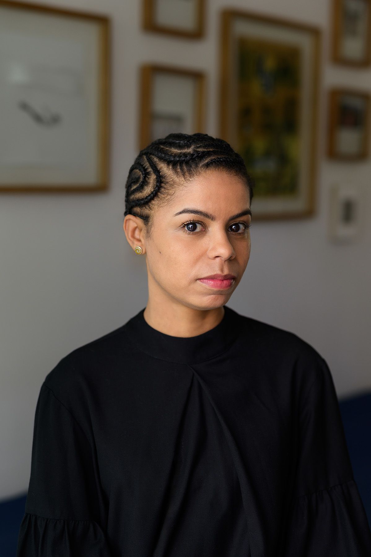 A woman with dark skin, dark eyes, and intricate spiral cornrows on her scalp wears a black high-neck top and looks at the camera.