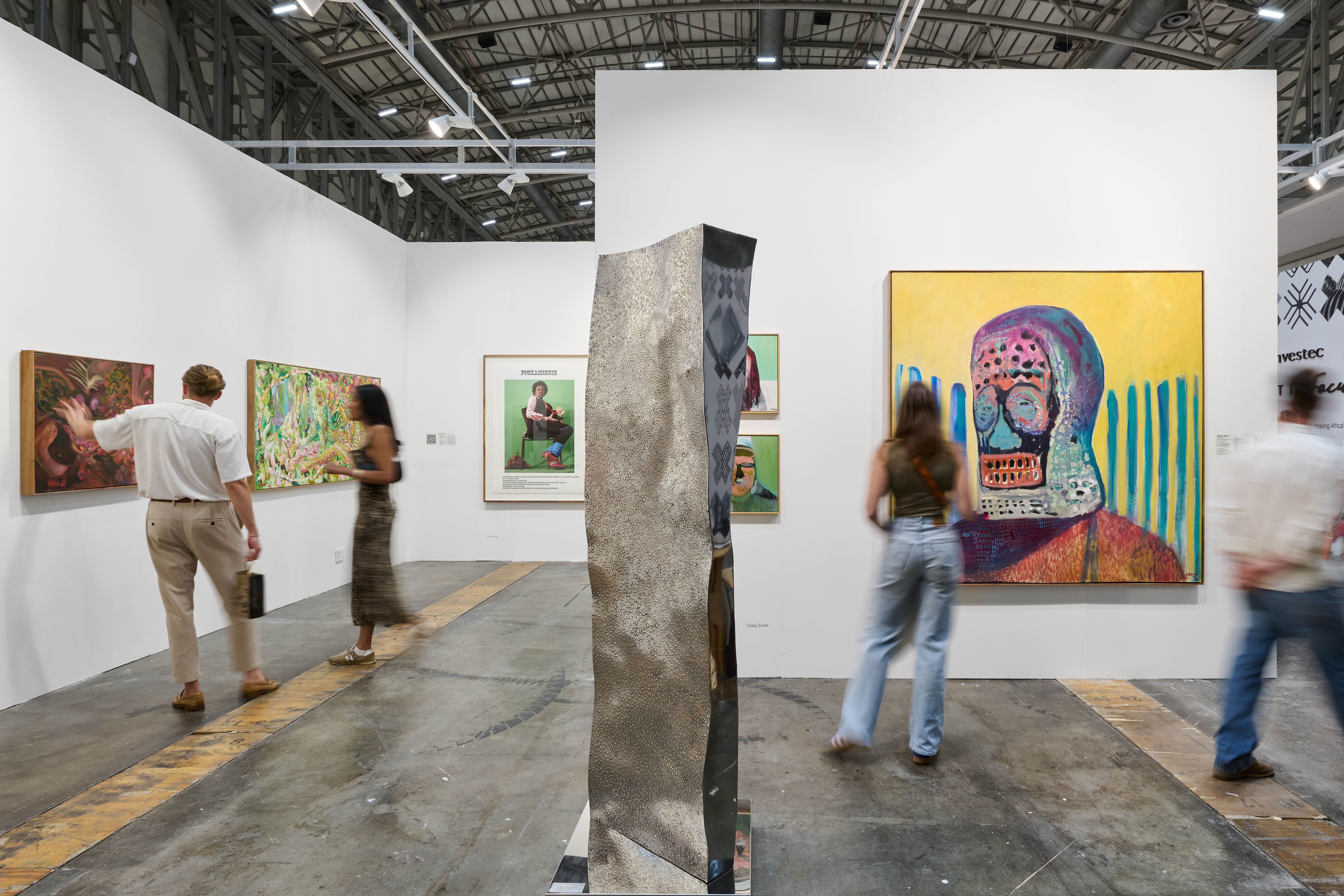 A bustling art fair with people viewing colorful paintings and a large, abstract sculpture.