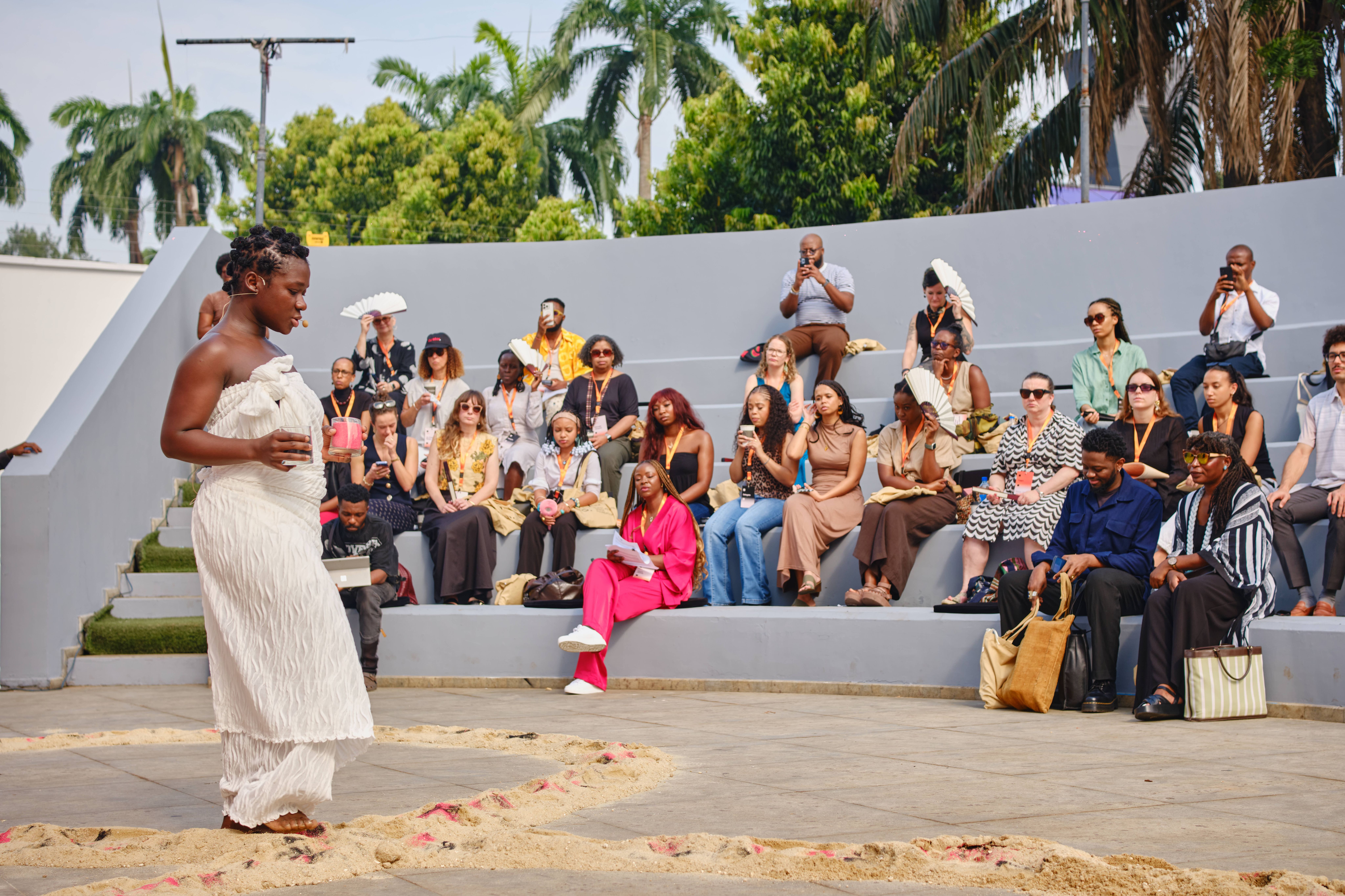 A performer in white stands in a sandy circle before an audience seated in an outdoor amphitheater.