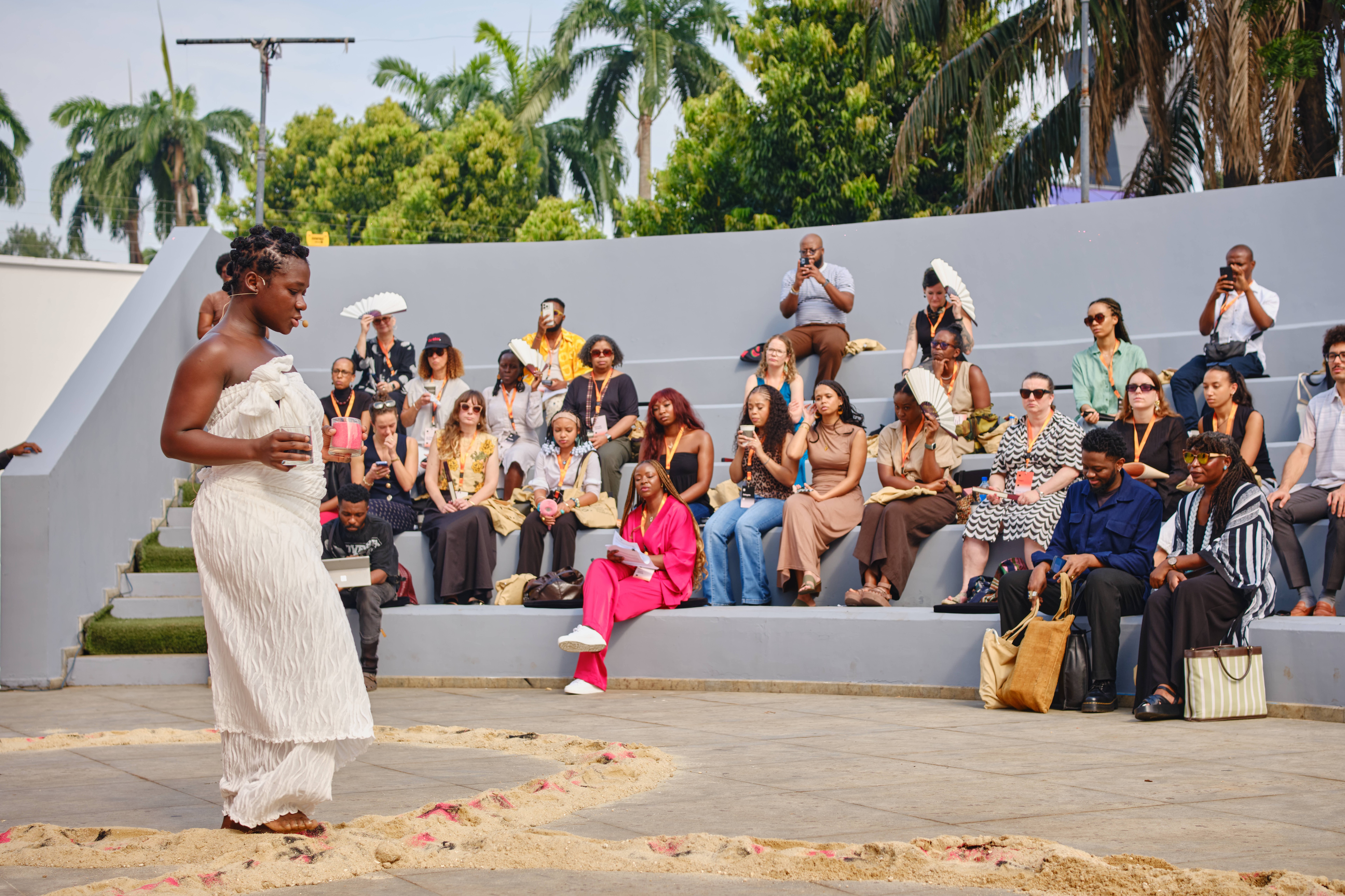 A performer in white stands in a sandy circle before an audience seated in an outdoor amphitheater.