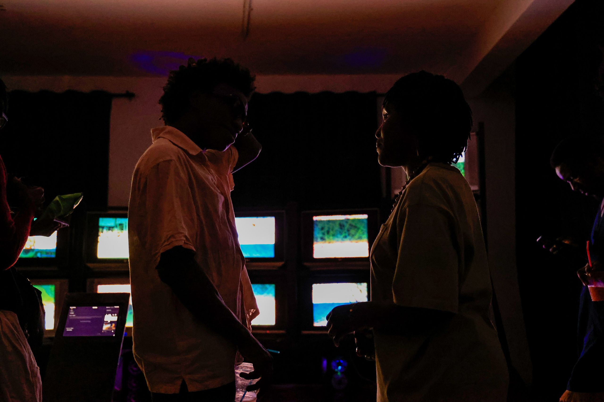Two people in silhouette stand in a dark room illuminated by several glowing screens.