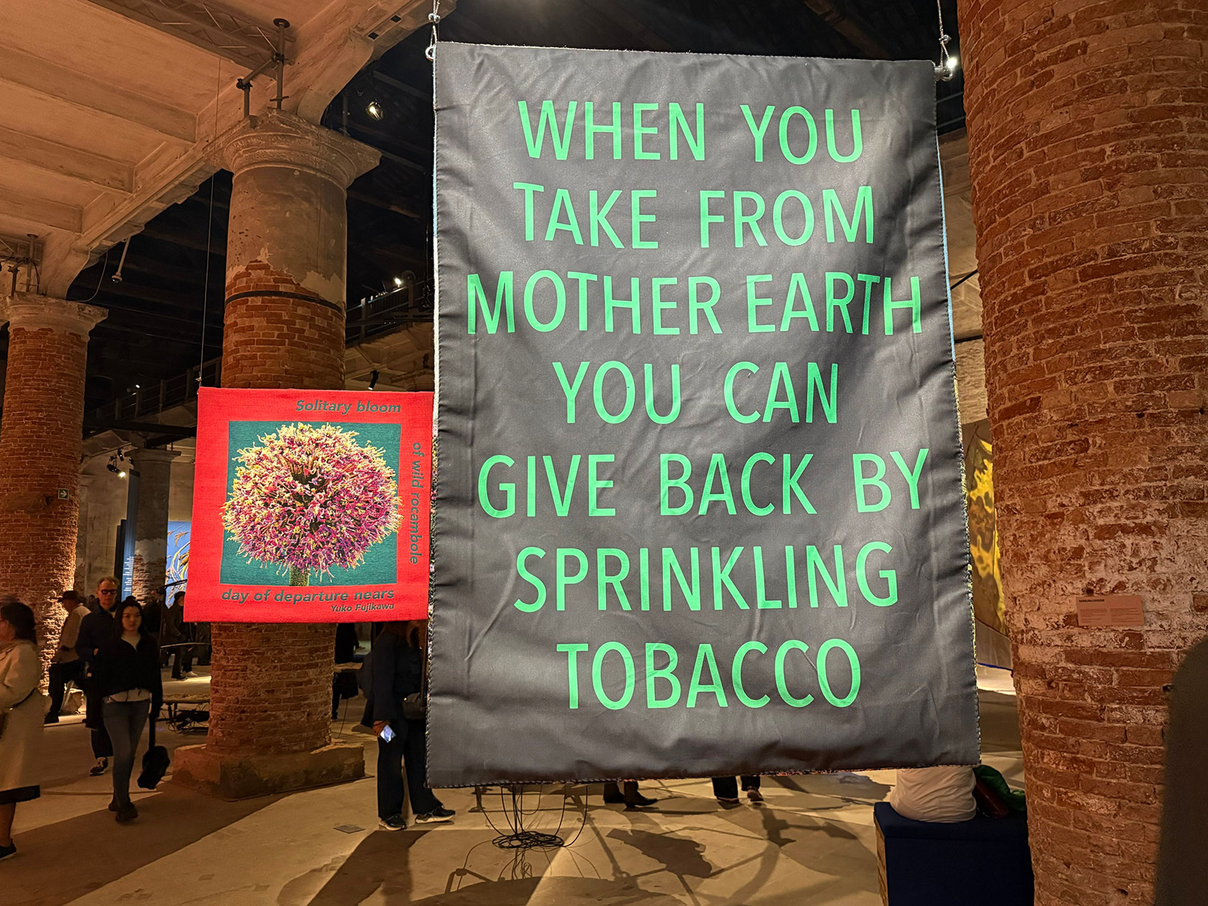 A gray banner with green text 'WHEN YOU TAKE FROM MOTHER EARTH YOU CAN GIVE BACK BY SPRINKLING TOBACCO' hangs in an art exhibit with a smaller red banner showing a pink flower and brick columns.