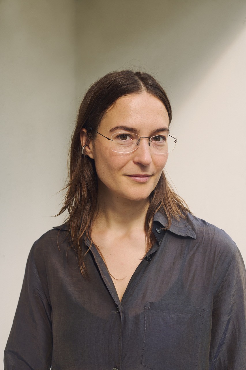 A woman with long brown hair and rimless glasses smiles slightly at the camera, wearing a sheer dark grey shirt.