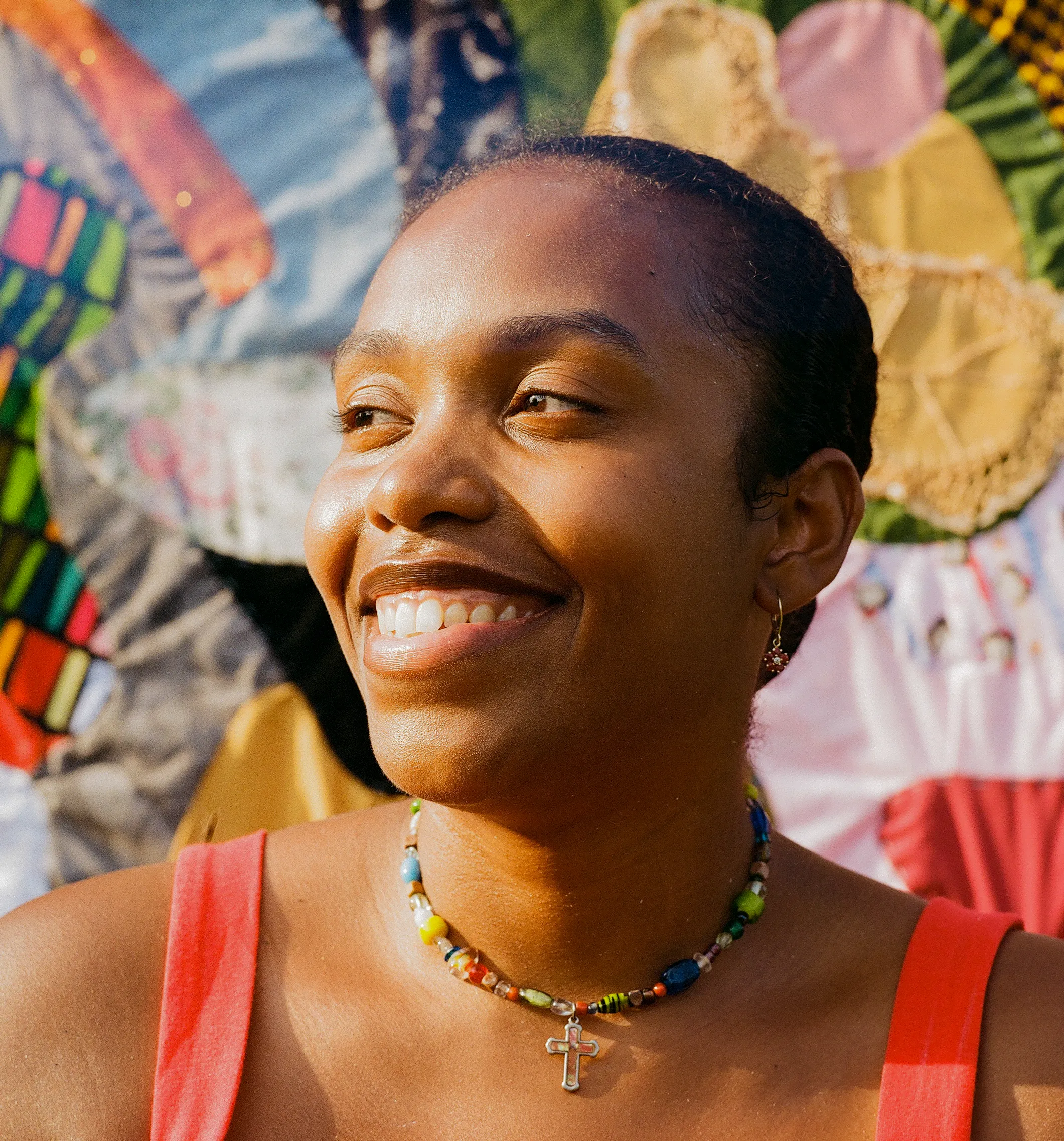 Close-up of a smiling Black woman looking left, wearing a colorful beaded necklace with a silver cross, against a vibrant, blurry textile.