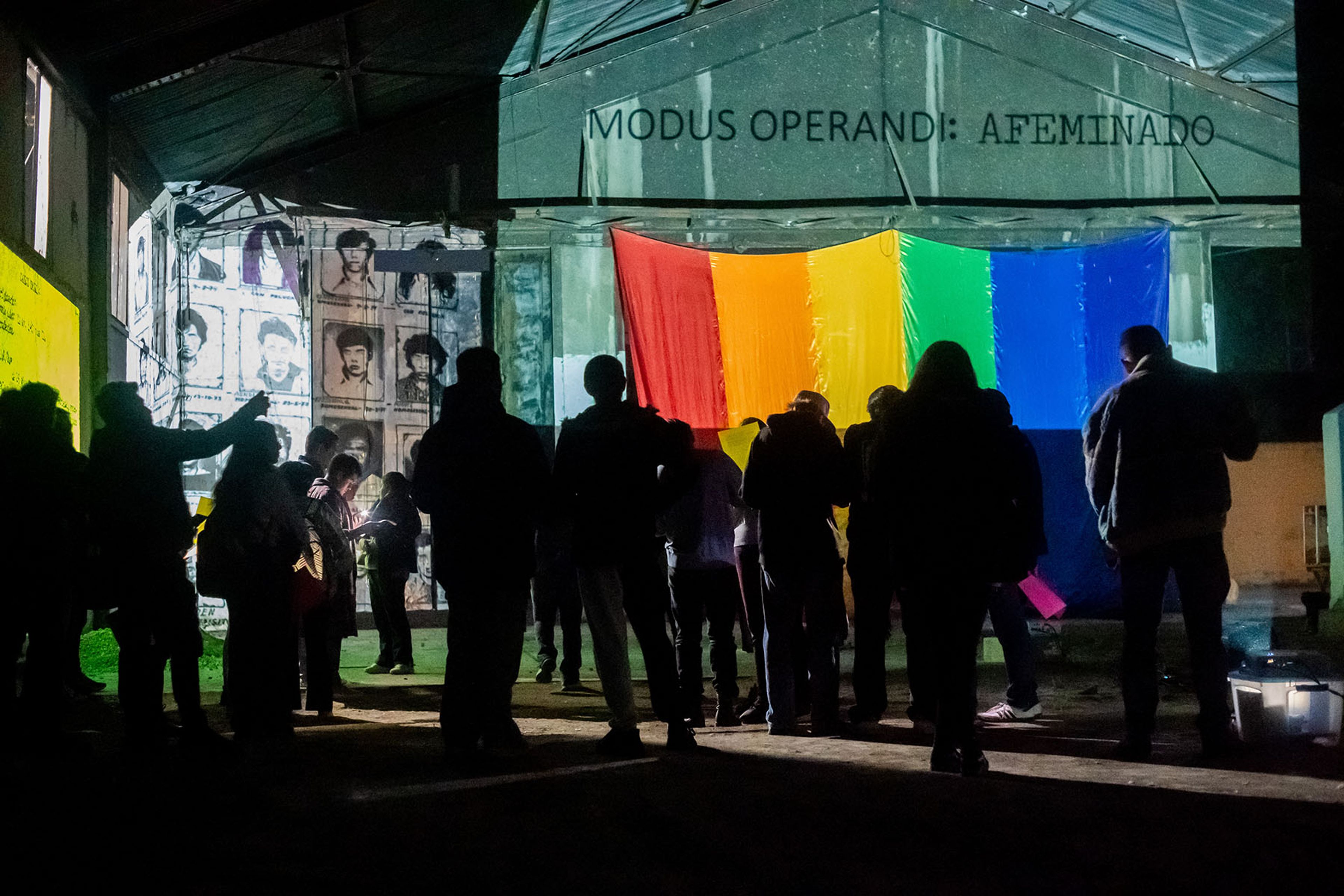 Silhouetted people view a building with a projected rainbow flag, portraits, and text "MODUS OPERANDI: AFEMINADO".