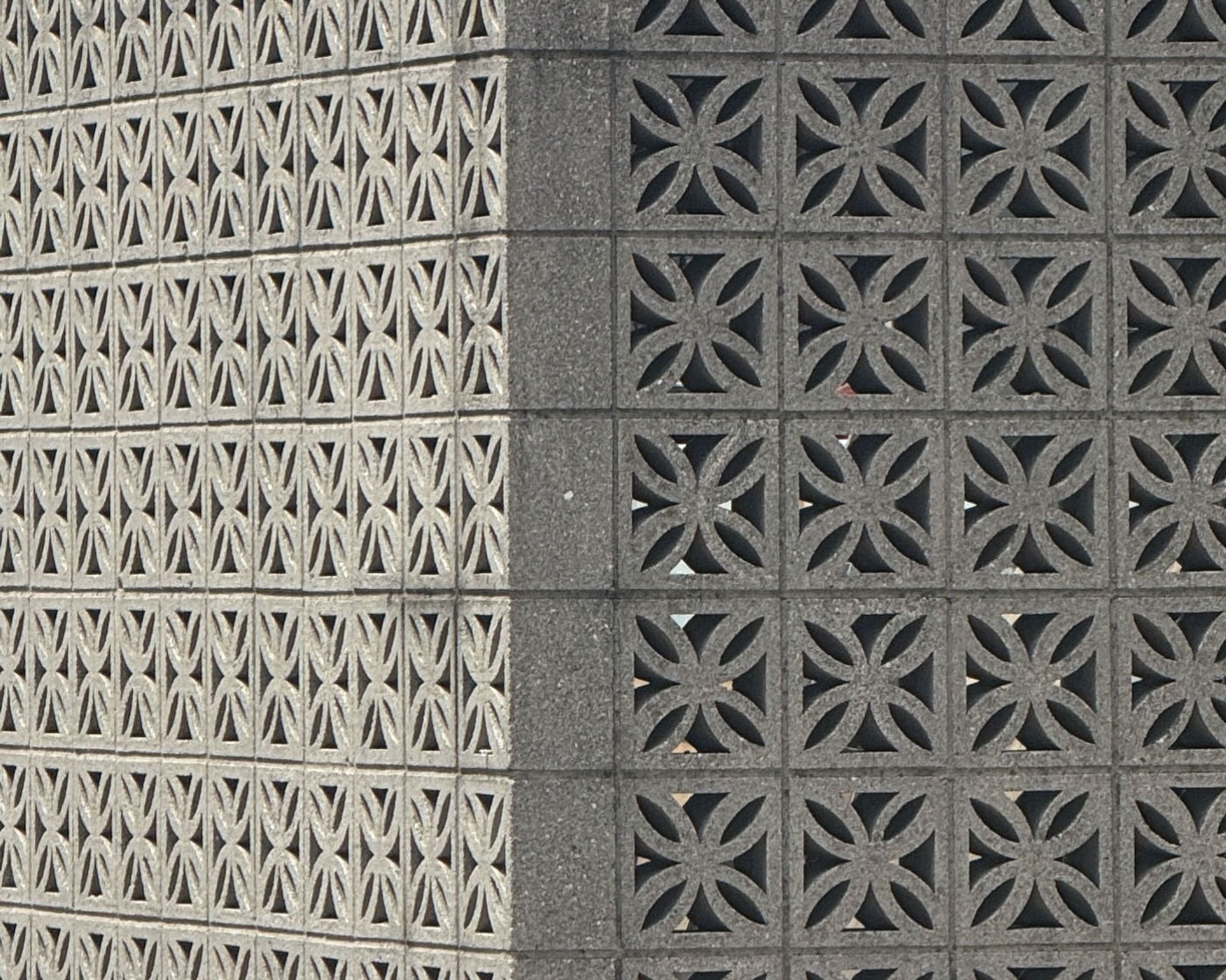 Corner of a decorative concrete block wall featuring two distinct geometric patterns: light grey hourglass shapes on the left, and dark grey four-petal flower shapes on the right.