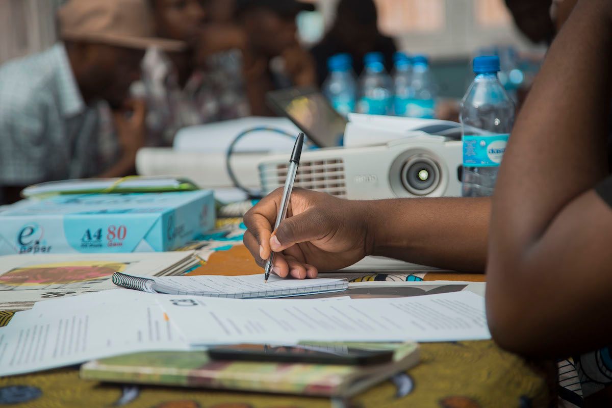 A person's hand writes in a notebook, with a projector and blurred people in the background.