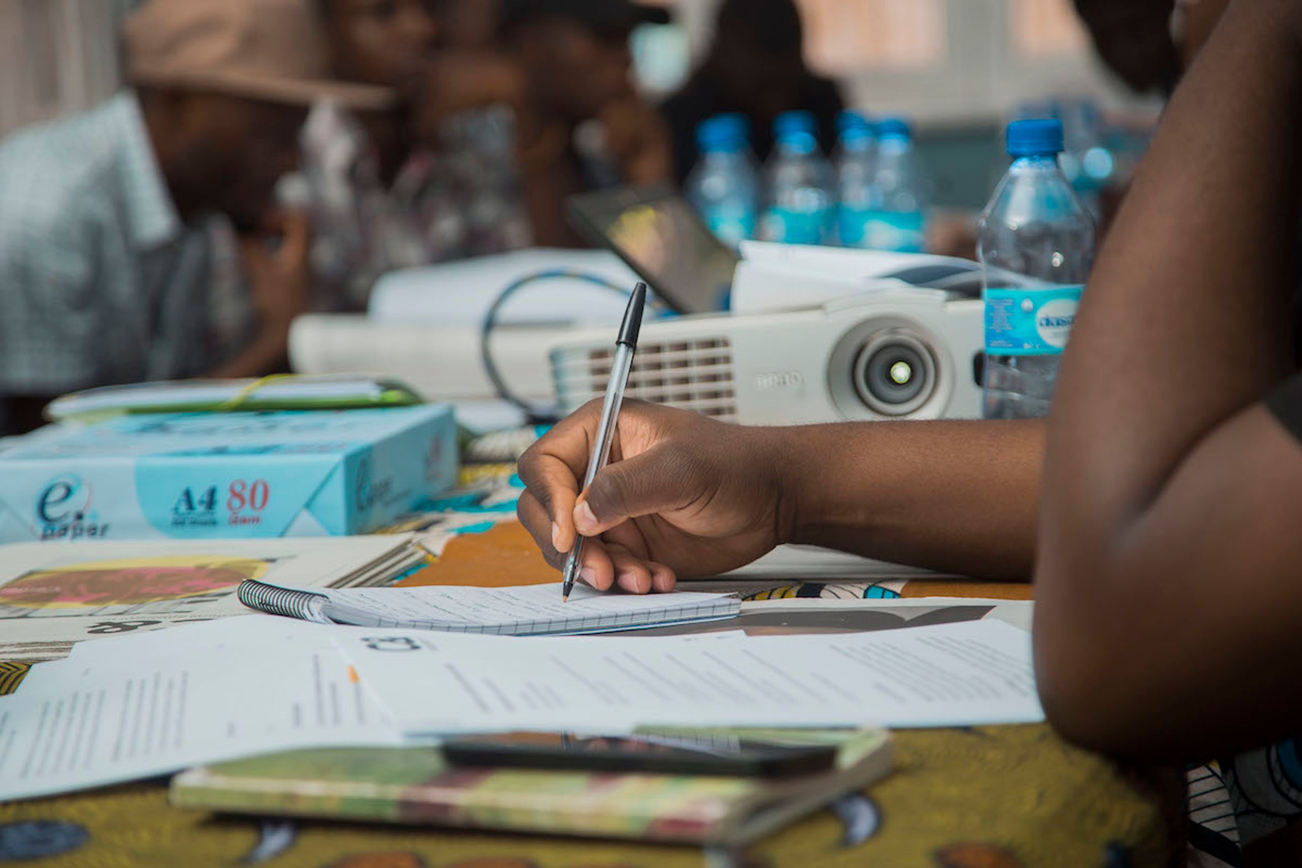 A person's hand writes in a notebook, with a projector and blurred people in the background.