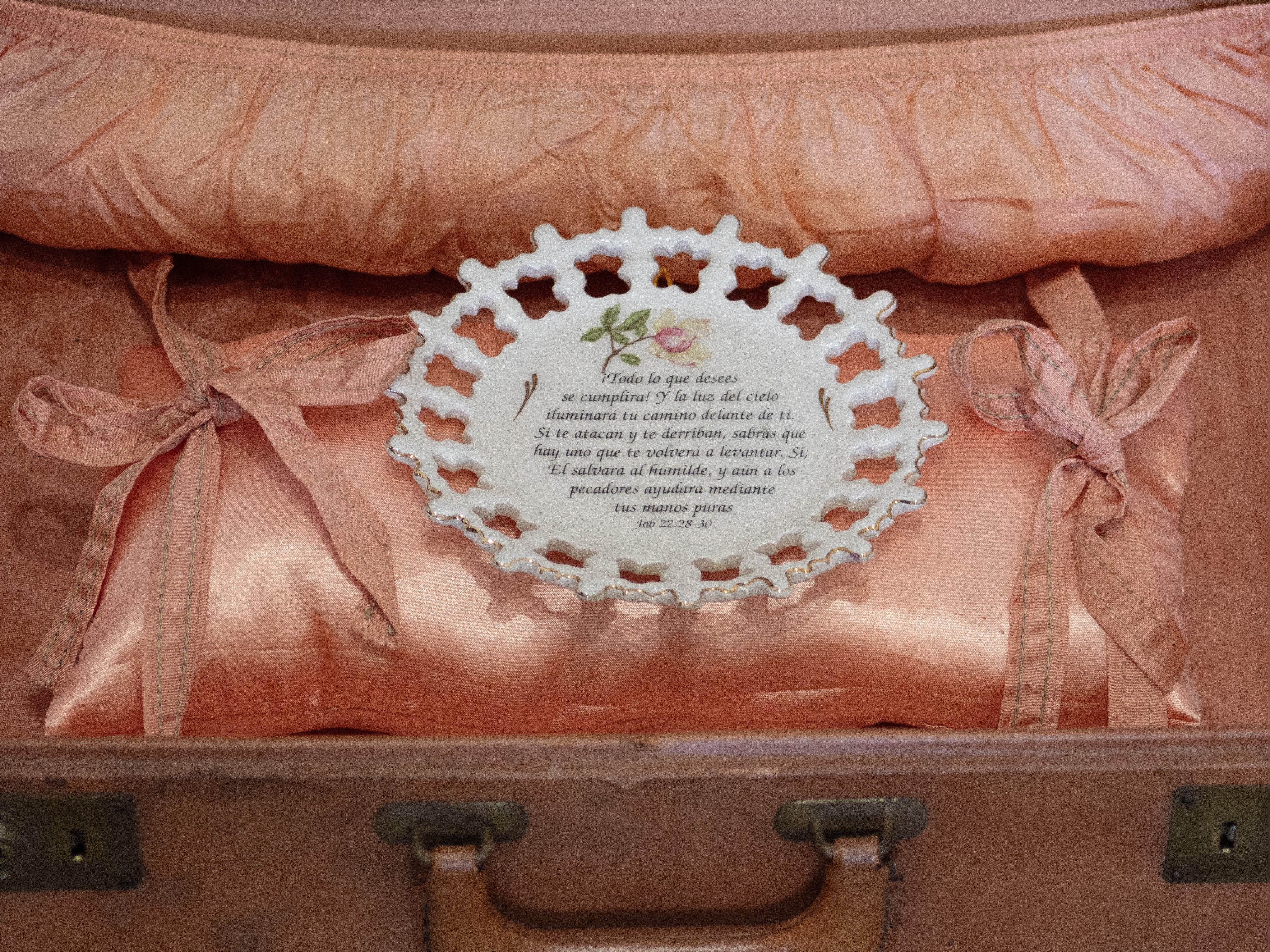 A white ceramic plate with a scalloped edge, rose design, and Spanish text from Job 22:28-30 lies on a peach satin pillow inside an open suitcase.