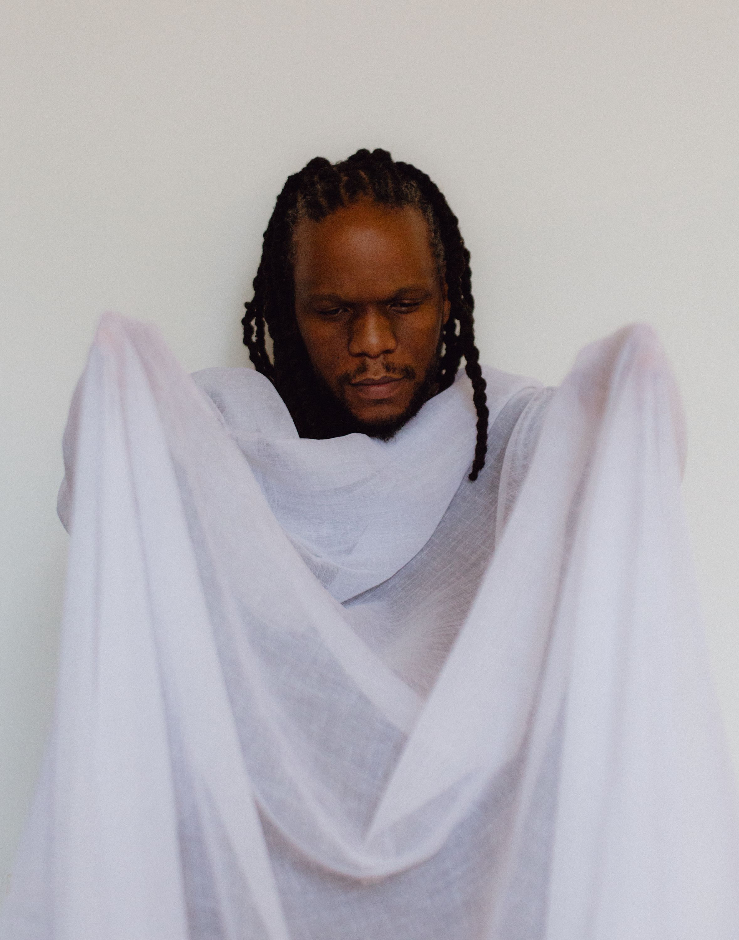A dark-skinned man with dreadlocks looks down, draped in white sheer fabric against a white background.