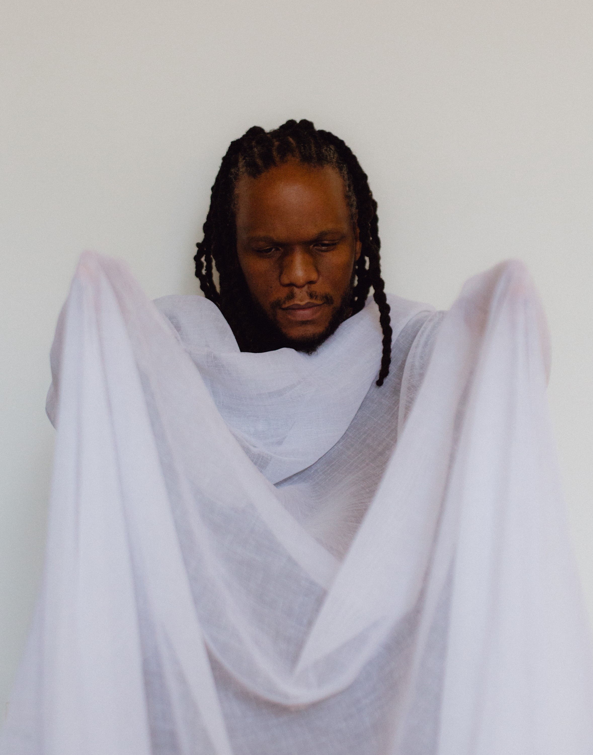 A dark-skinned man with dreadlocks looks down, draped in white sheer fabric against a white background.