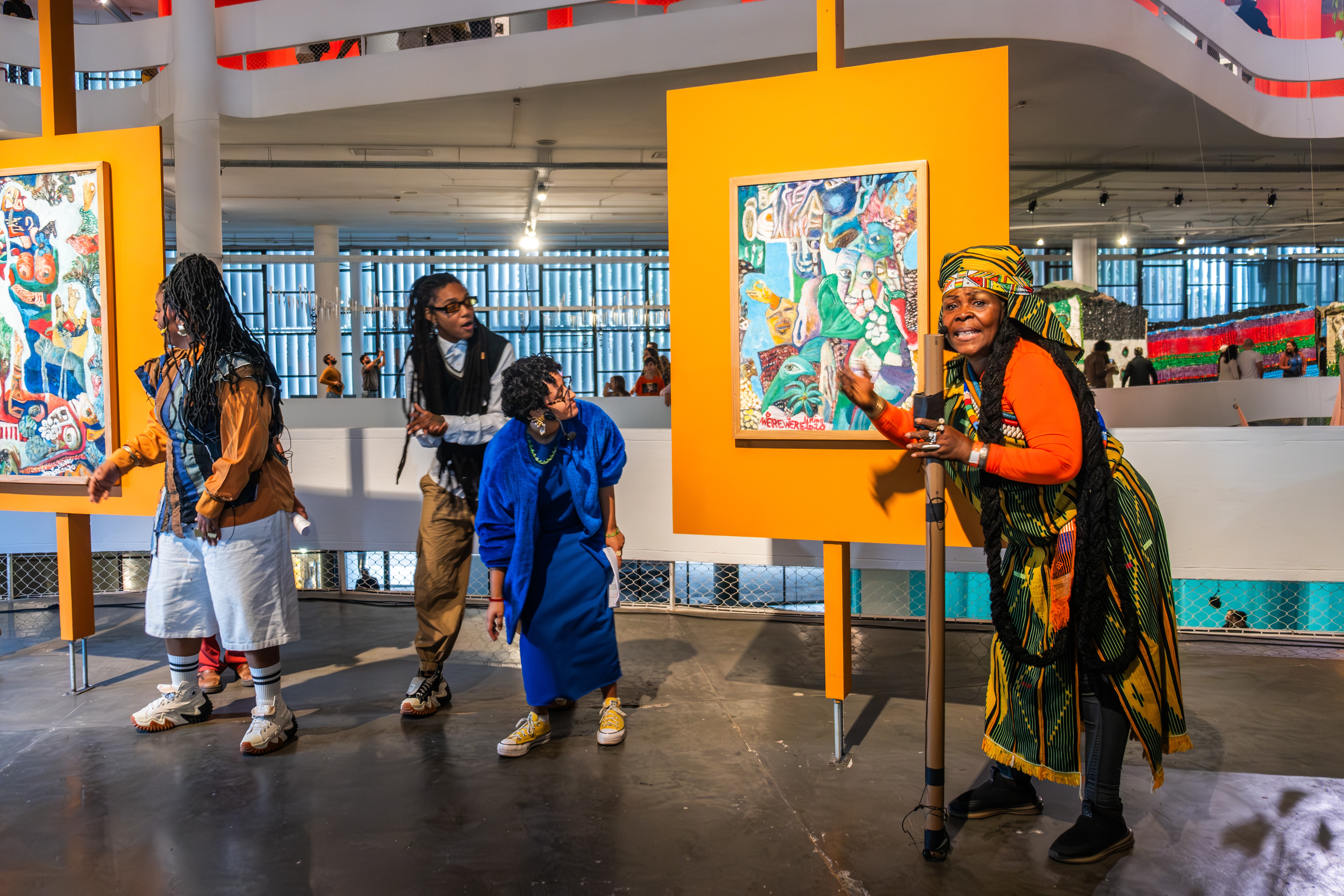 Four people in an art gallery, one smiling woman in traditional attire gestures toward a colorful painting on a bright orange easel.