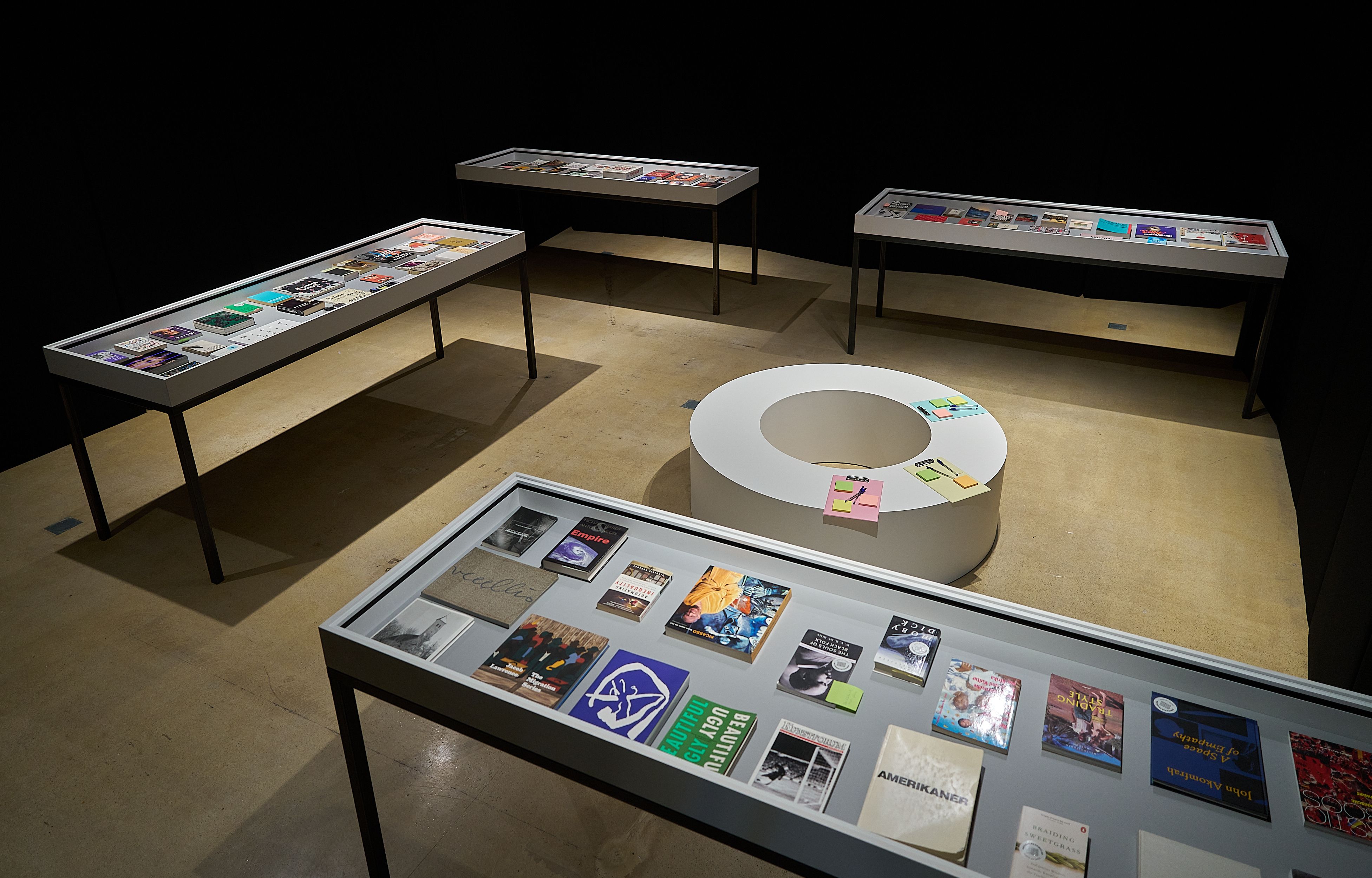 An exhibition space with multiple glass display cases filled with books and magazines, and a white circular table with colorful notes.