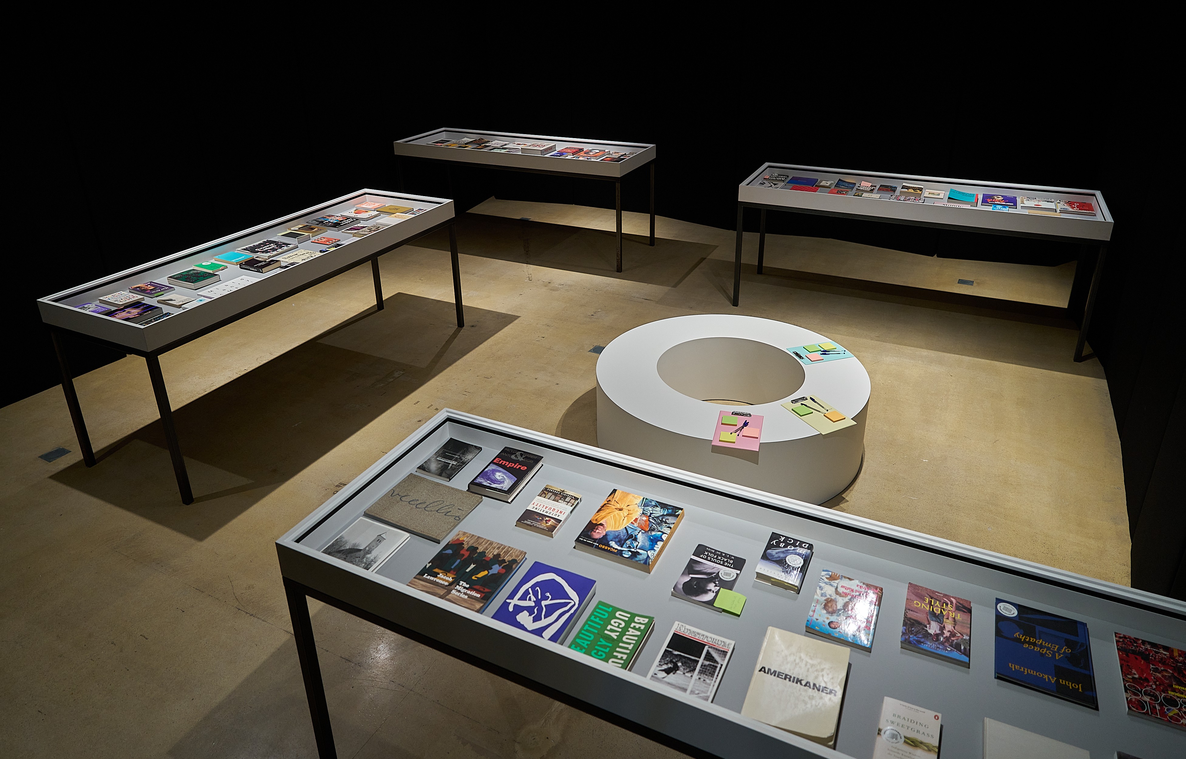 An exhibition space with multiple glass display cases filled with books and magazines, and a white circular table with colorful notes.