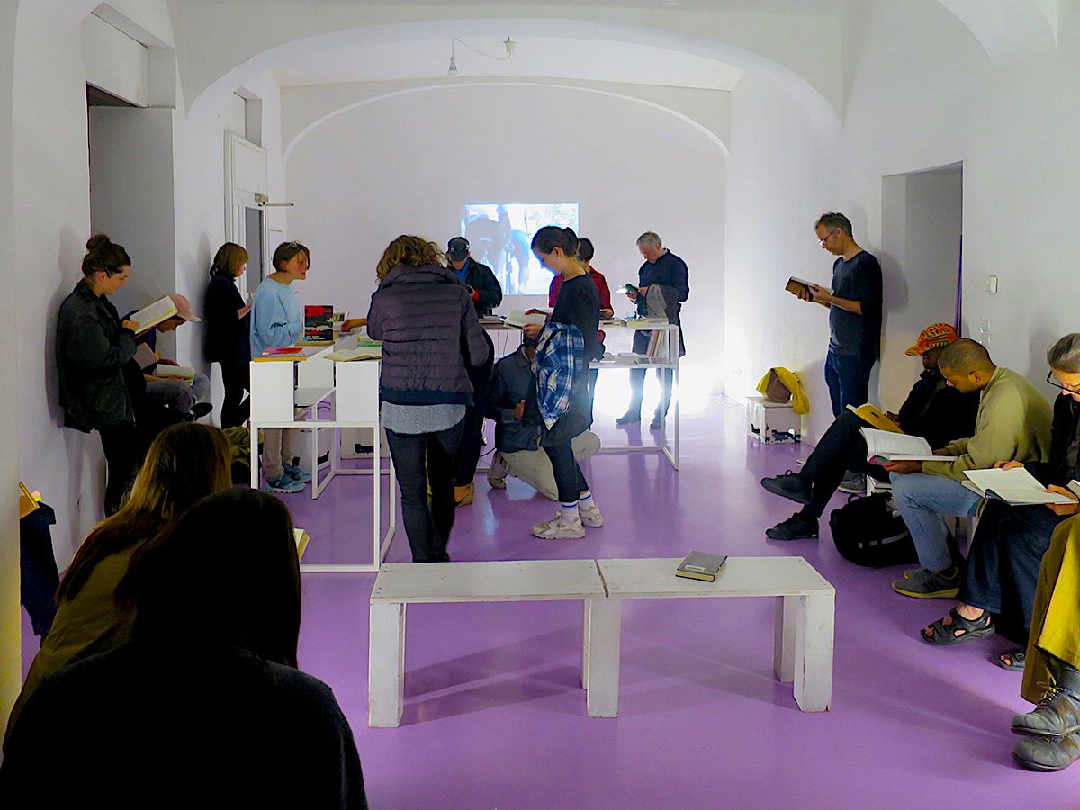 A group of people reading and looking at books in a bright room with white walls and a purple floor, with a projection on the far wall.