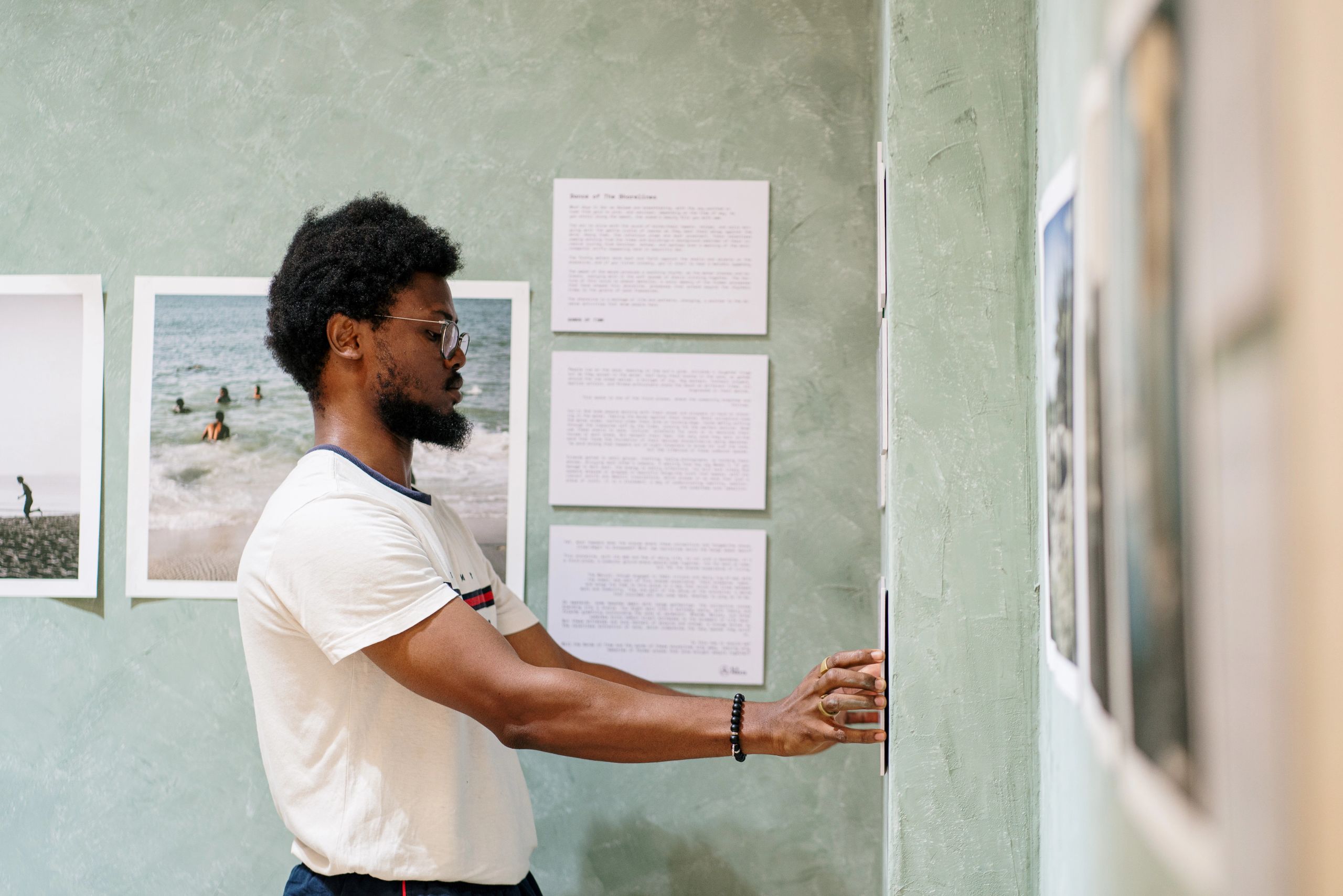 A man with curly hair and a beard adjusts a framed photograph on a green wall in an exhibition.