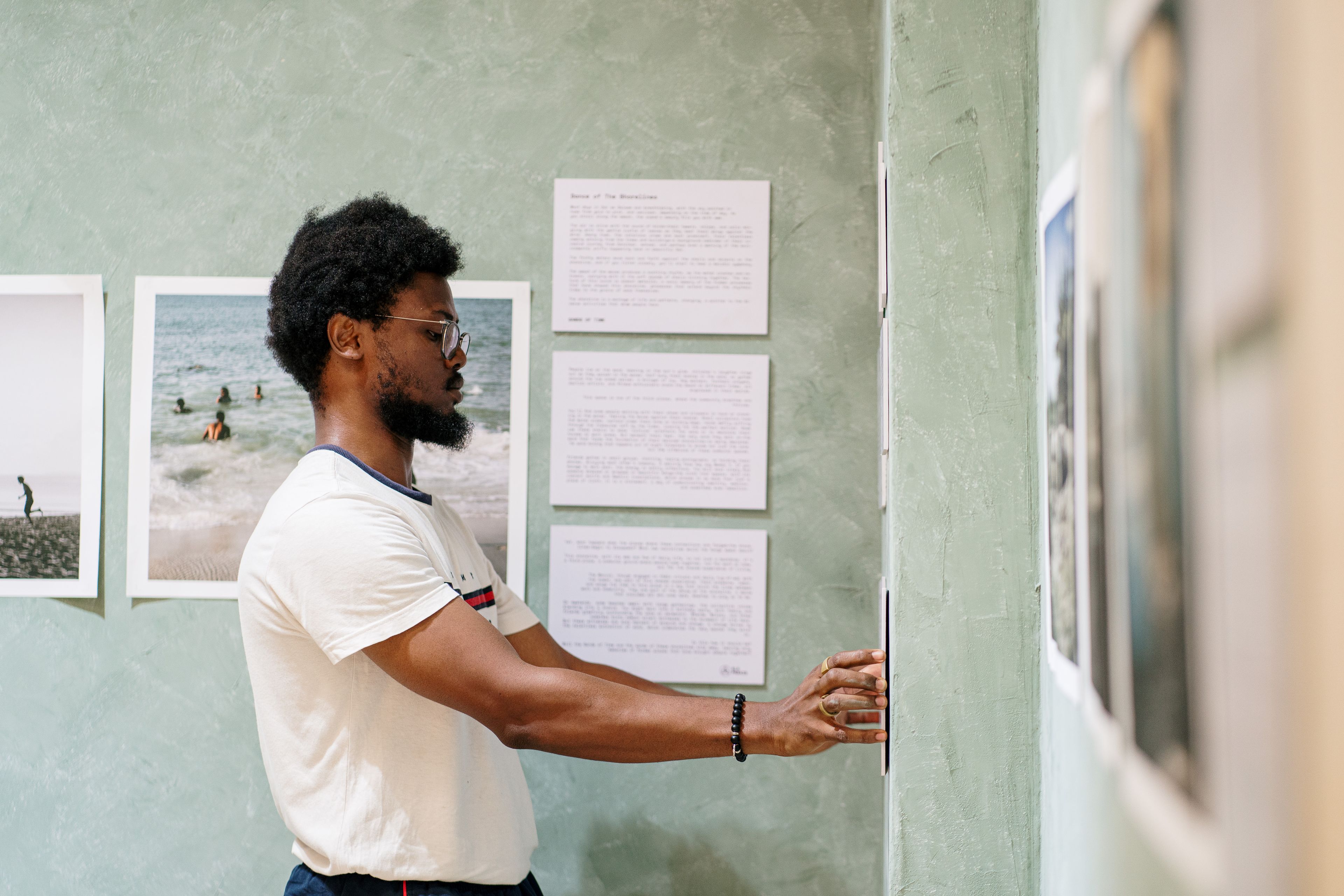 A man with curly hair and a beard adjusts a framed photograph on a green wall in an exhibition.