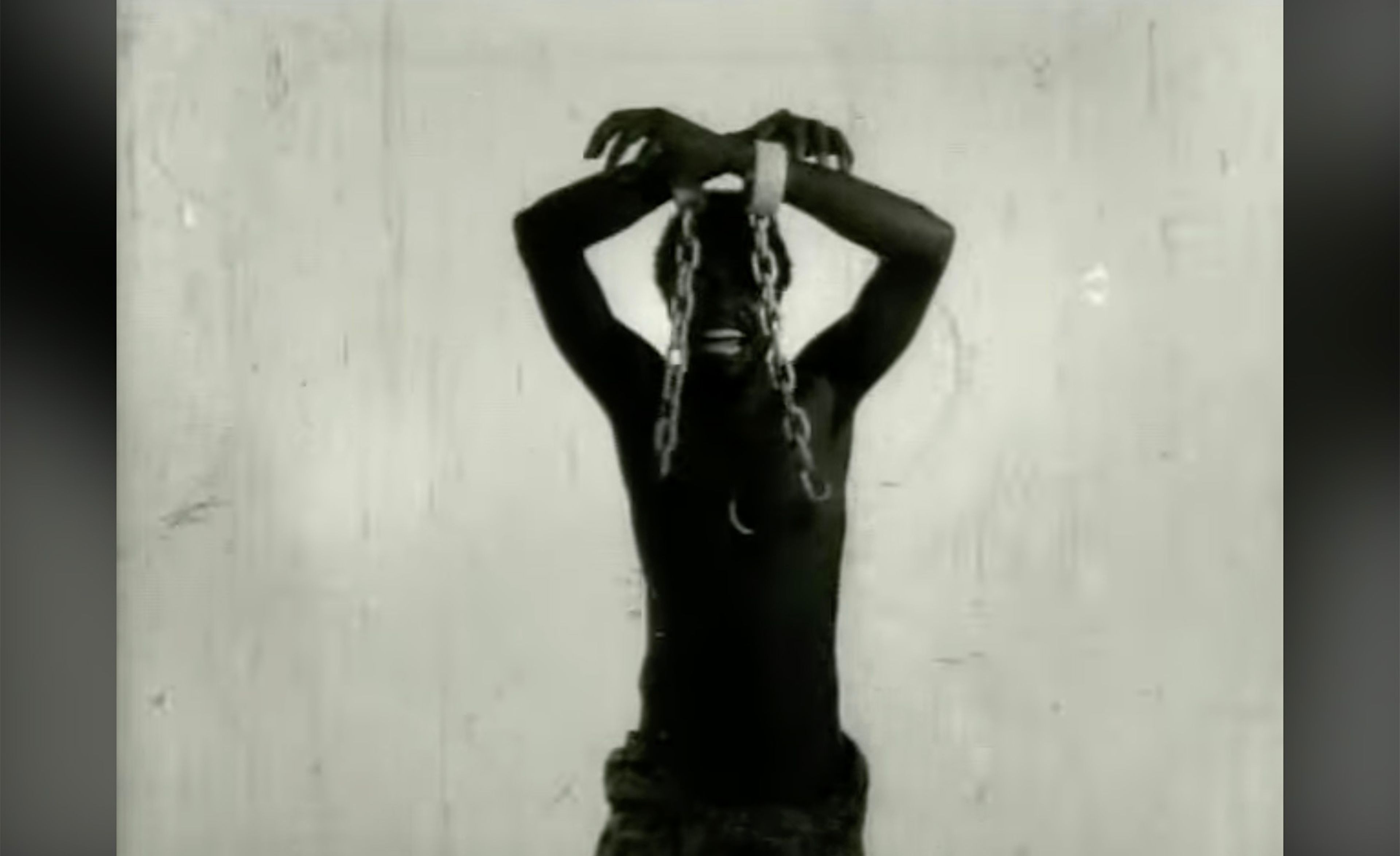 Black and white image of a shirtless man with dark skin, his chained hands raised overhead, grimacing.