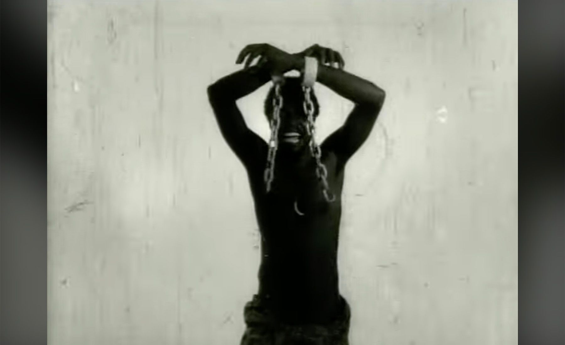 Black and white image of a shirtless man with dark skin, his chained hands raised overhead, grimacing.