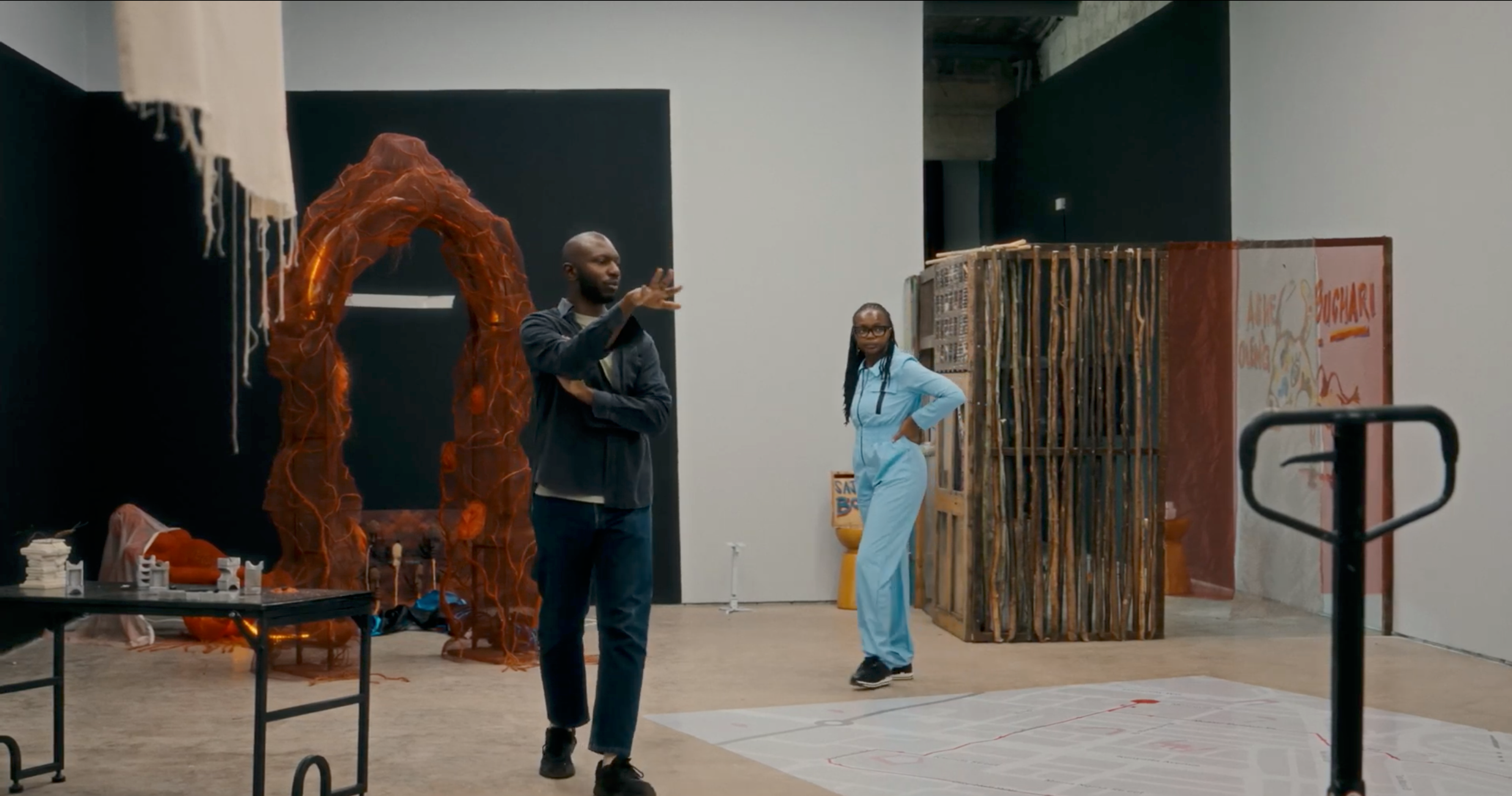 A man gestures near a large reddish archway sculpture, with a woman in a blue jumpsuit standing in the background of an art gallery.