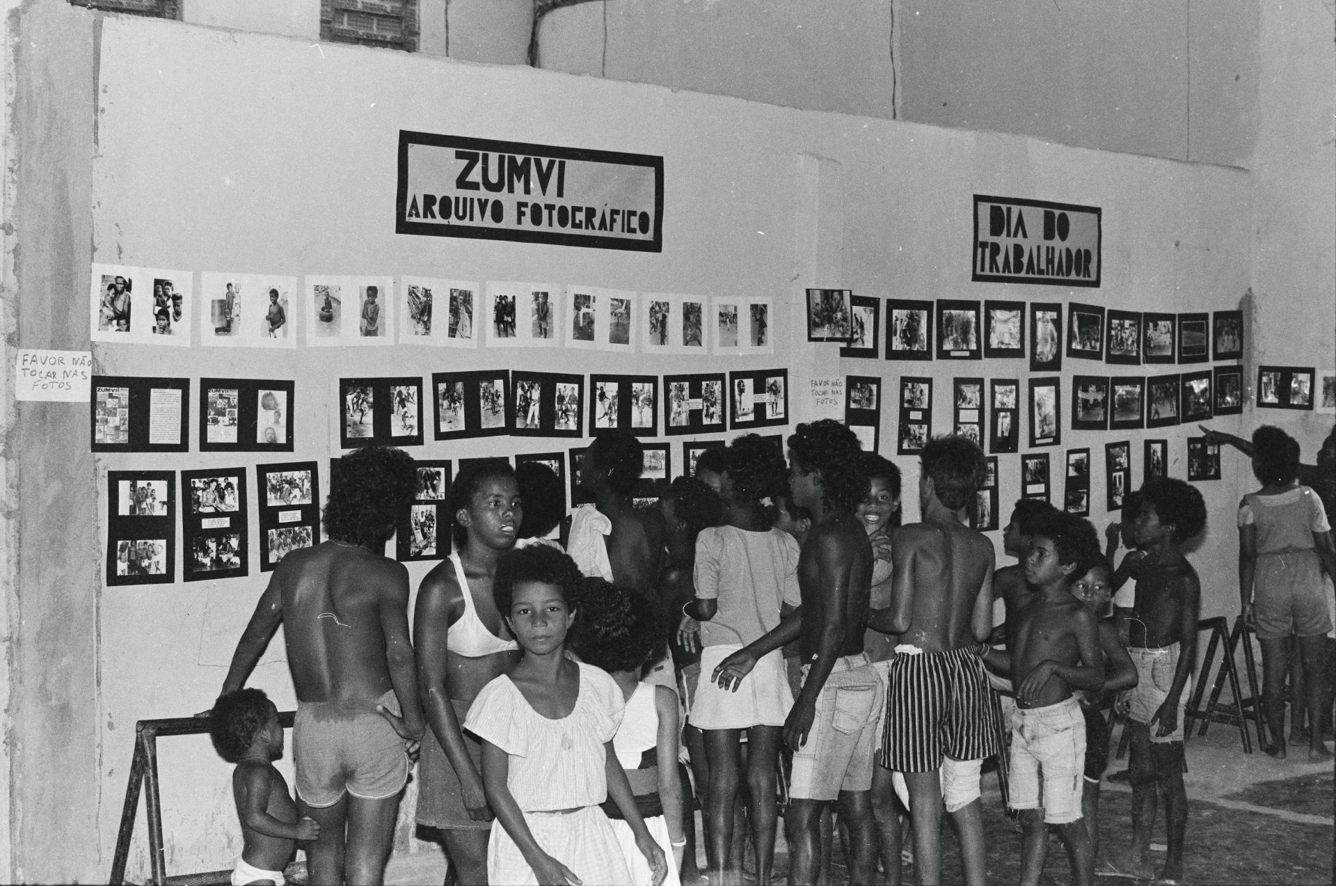 Black and white photo of a group of children viewing an exhibition of numerous framed photos on a wall, with signs for "Zumvi Arquivo Fotografico" and "Dia do Trabalhador".