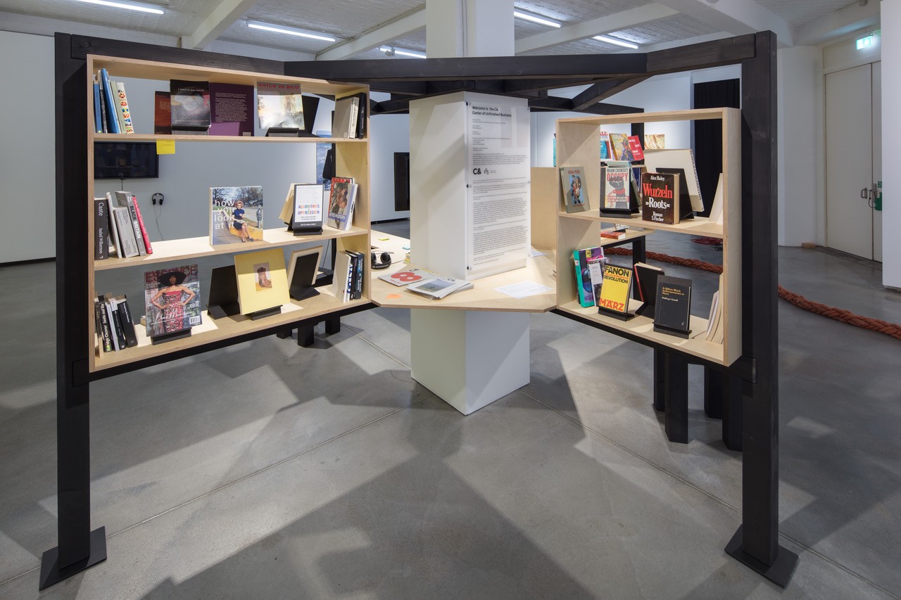 A multi-sided dark metal and light wood bookshelf display filled with books and a central informational panel in a gallery.