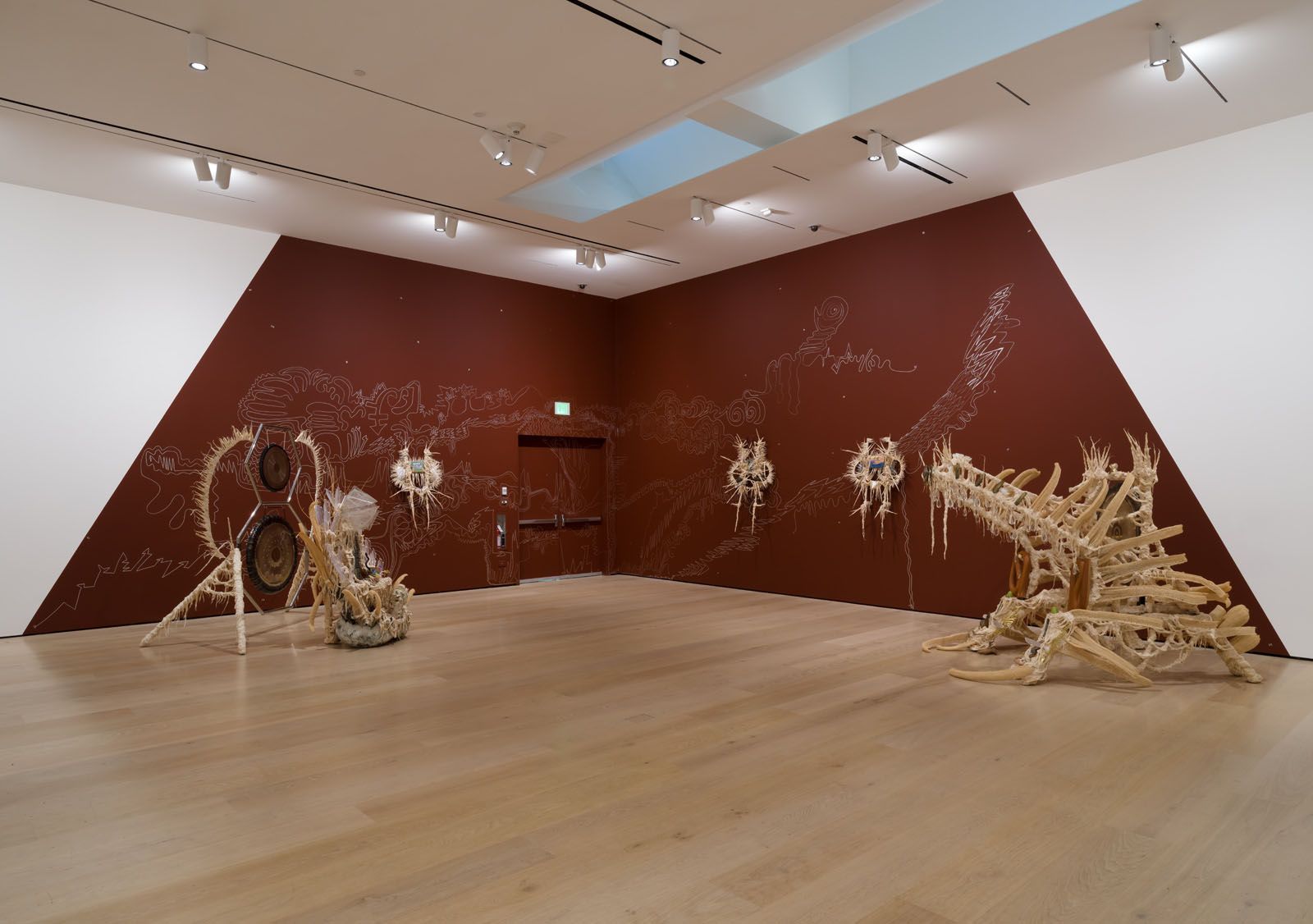 An art gallery with a reddish-brown wall featuring white abstract line drawings, a light wood floor, and multiple sculptures made of natural materials: a large circular piece on the left, spiky wall-mounted forms, and an intricate, bone-like structure on the right.