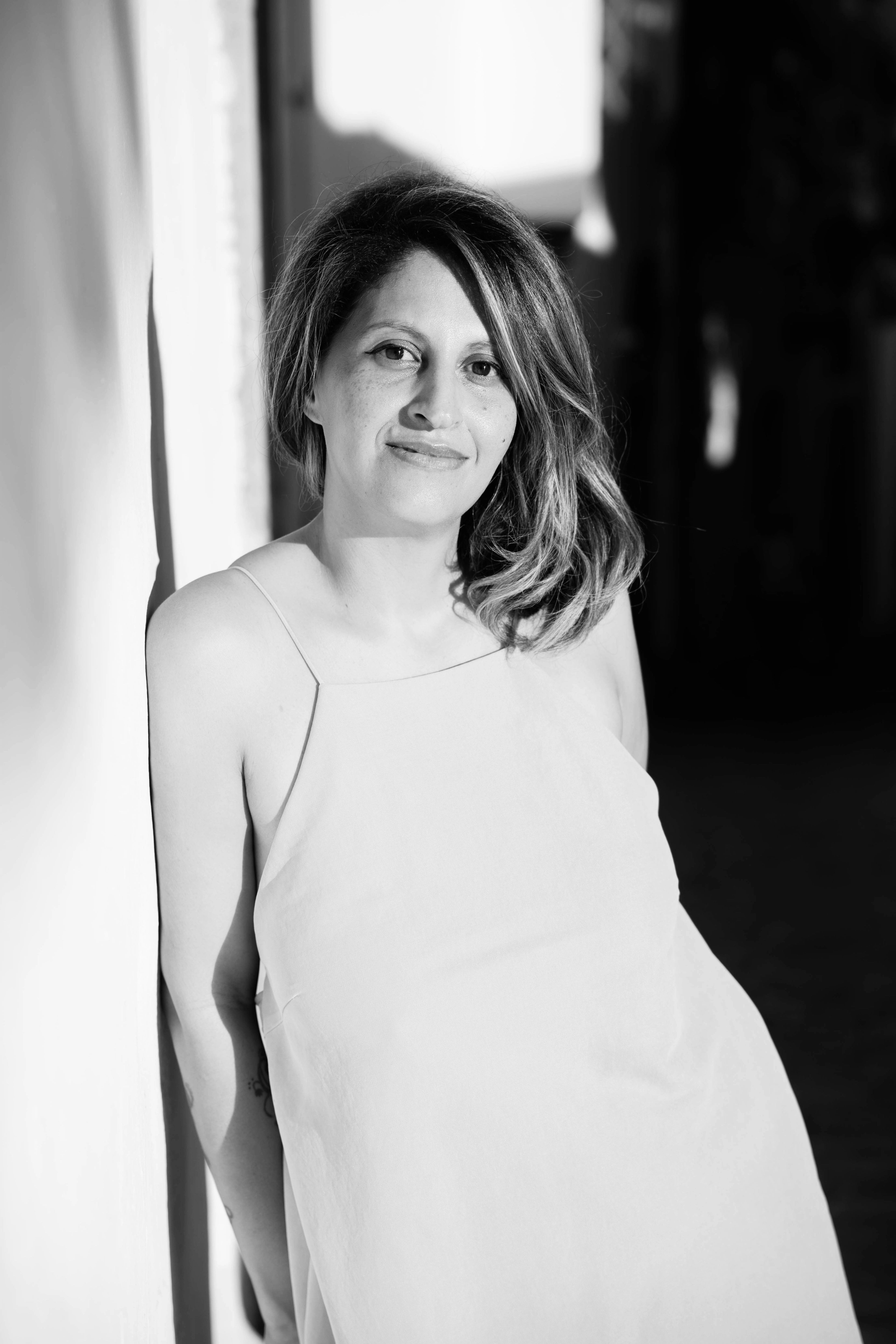 A smiling woman with wavy hair and freckles leans against a wall in a black and white photo.