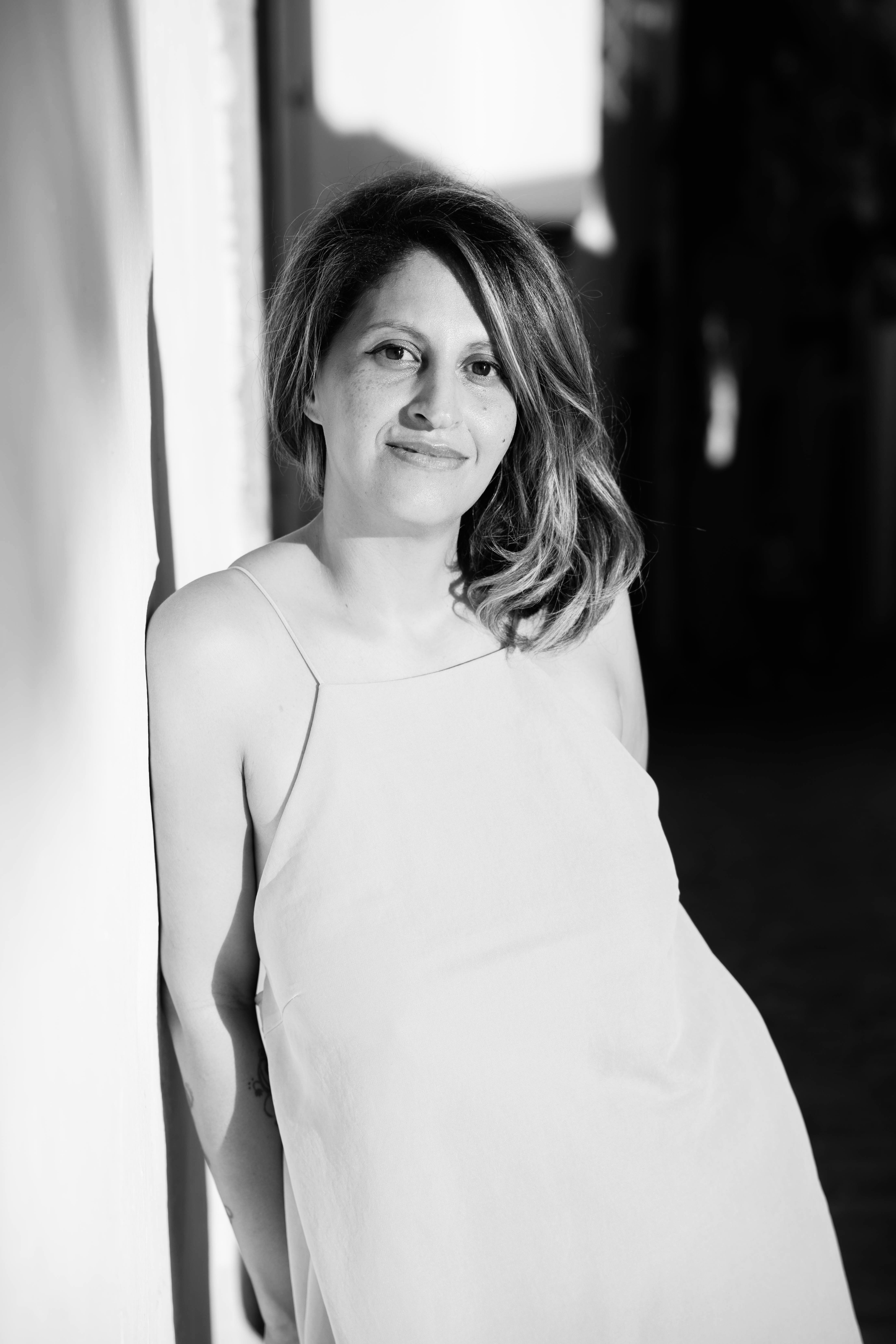A smiling woman with wavy hair and freckles leans against a wall in a black and white photo.