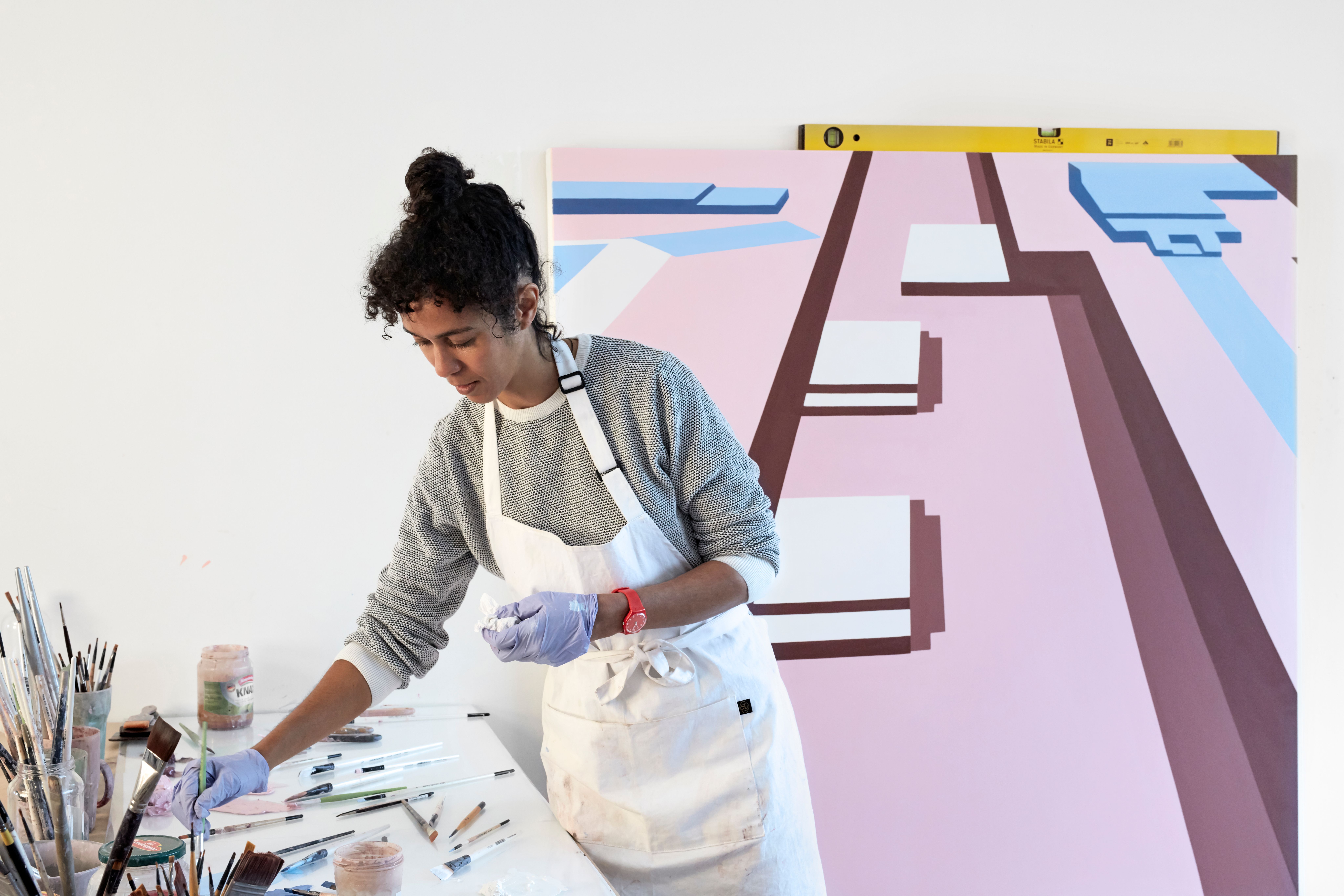 A woman artist in an apron and gloves reaches for supplies on a table, with a large abstract pink and blue painting in the background.