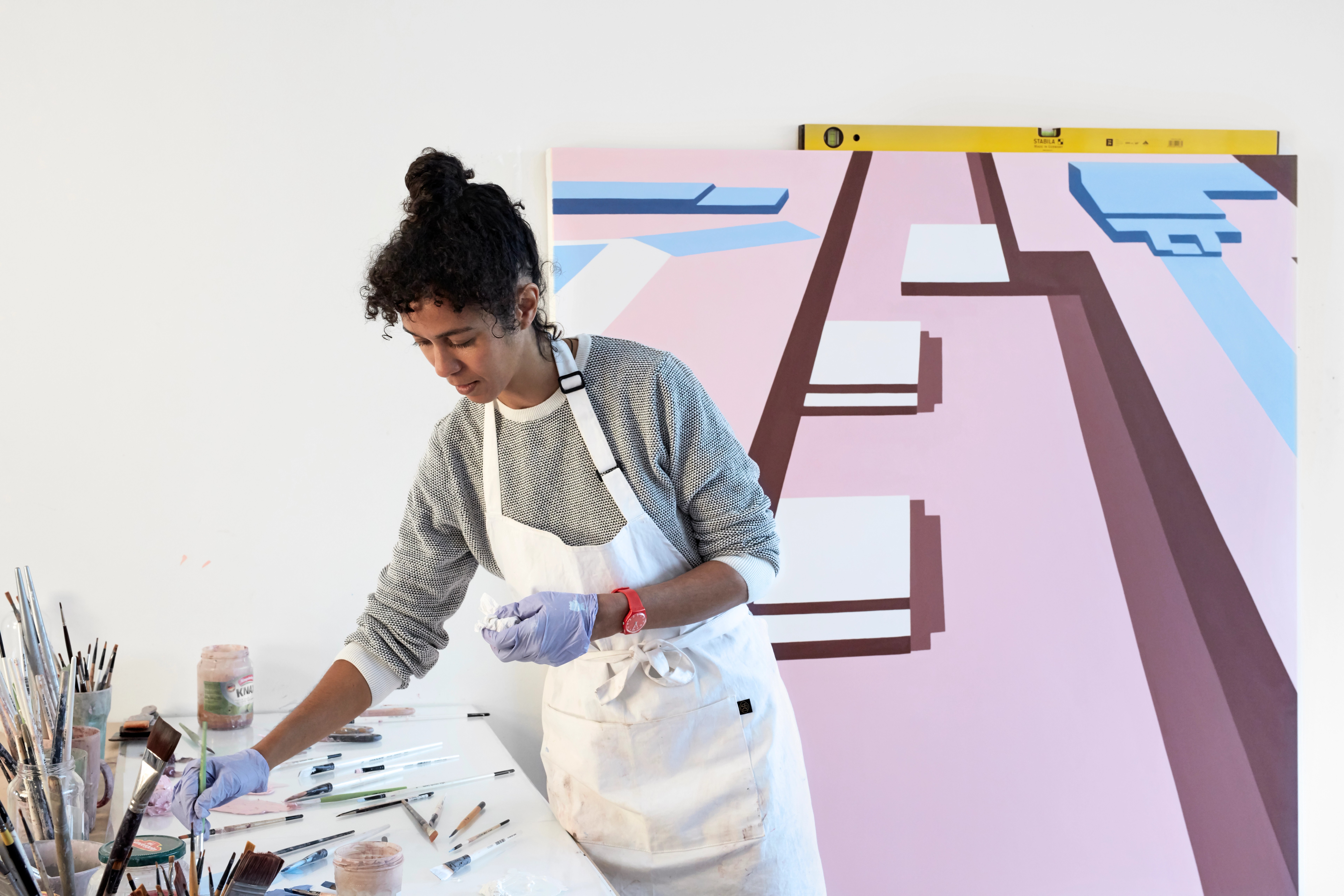 A woman artist in an apron and gloves reaches for supplies on a table, with a large abstract pink and blue painting in the background.