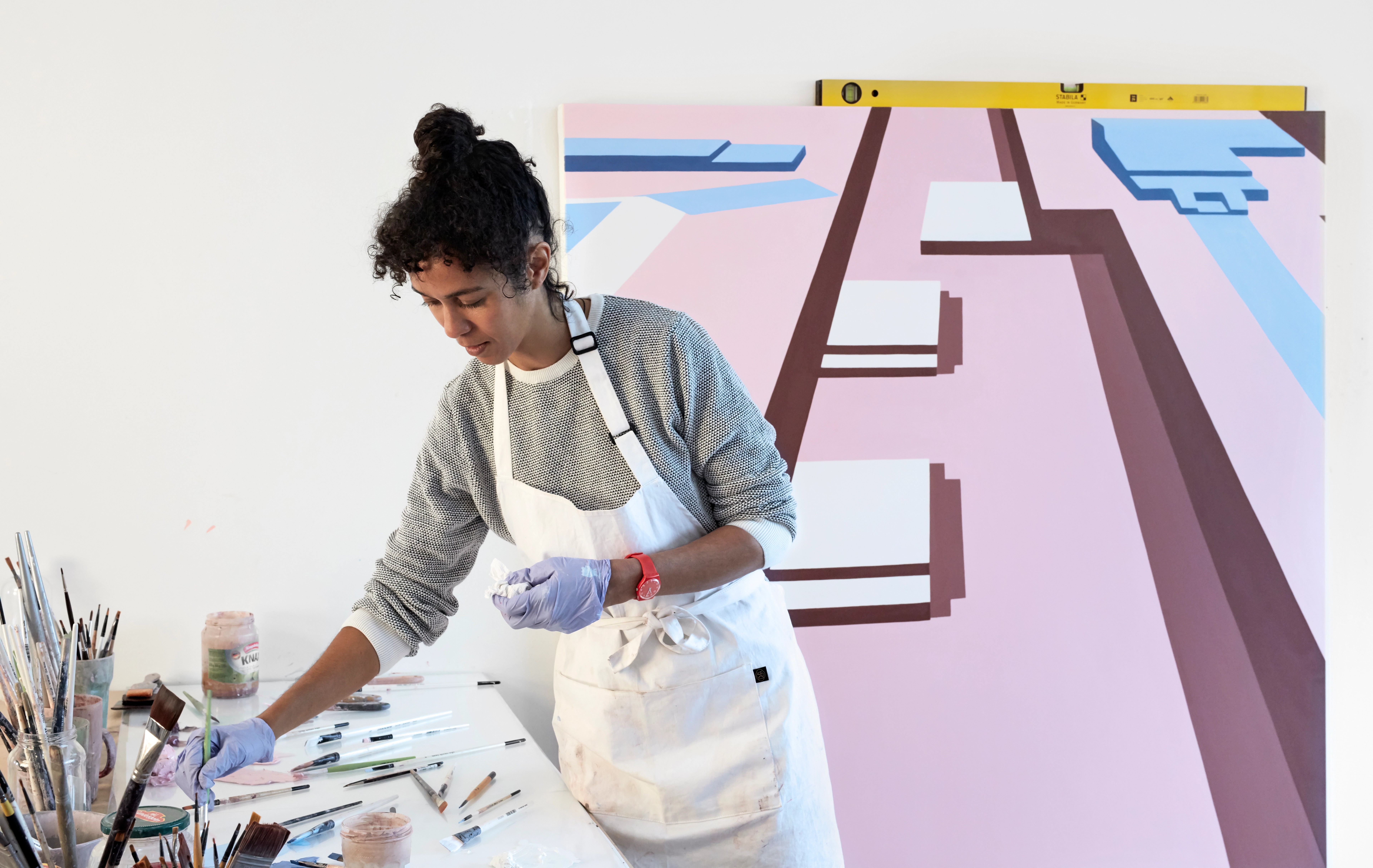 A female artist wearing an apron and gloves works at a table with art supplies, with a large abstract geometric painting behind her.
